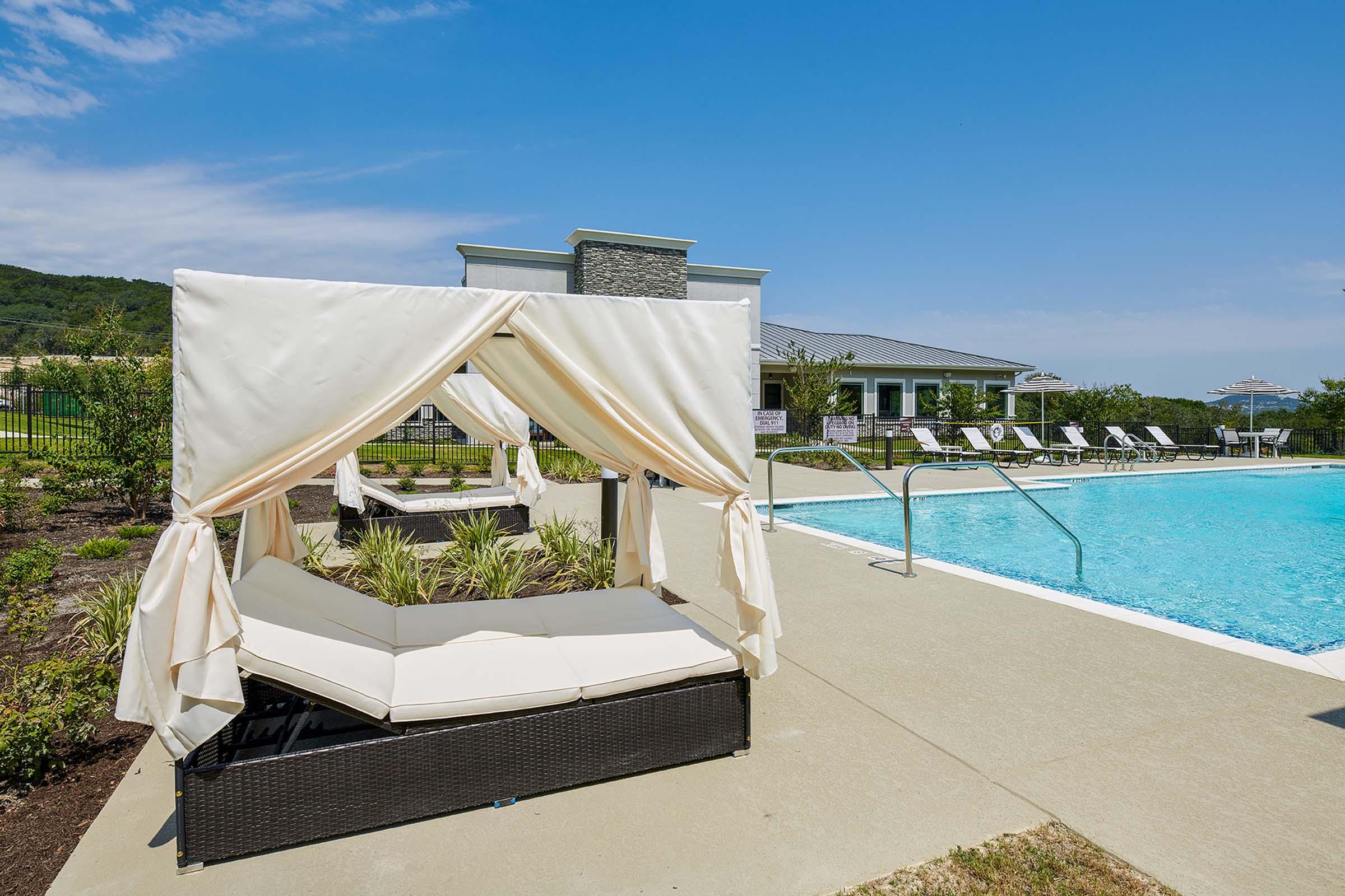 A serene outdoor pool area featuring a cozy cabana with comfortable seating, surrounded by lush greenery. In the background, there are lounge chairs and a modern building, with clear blue sky and distant hills visible, creating a relaxing atmosphere.