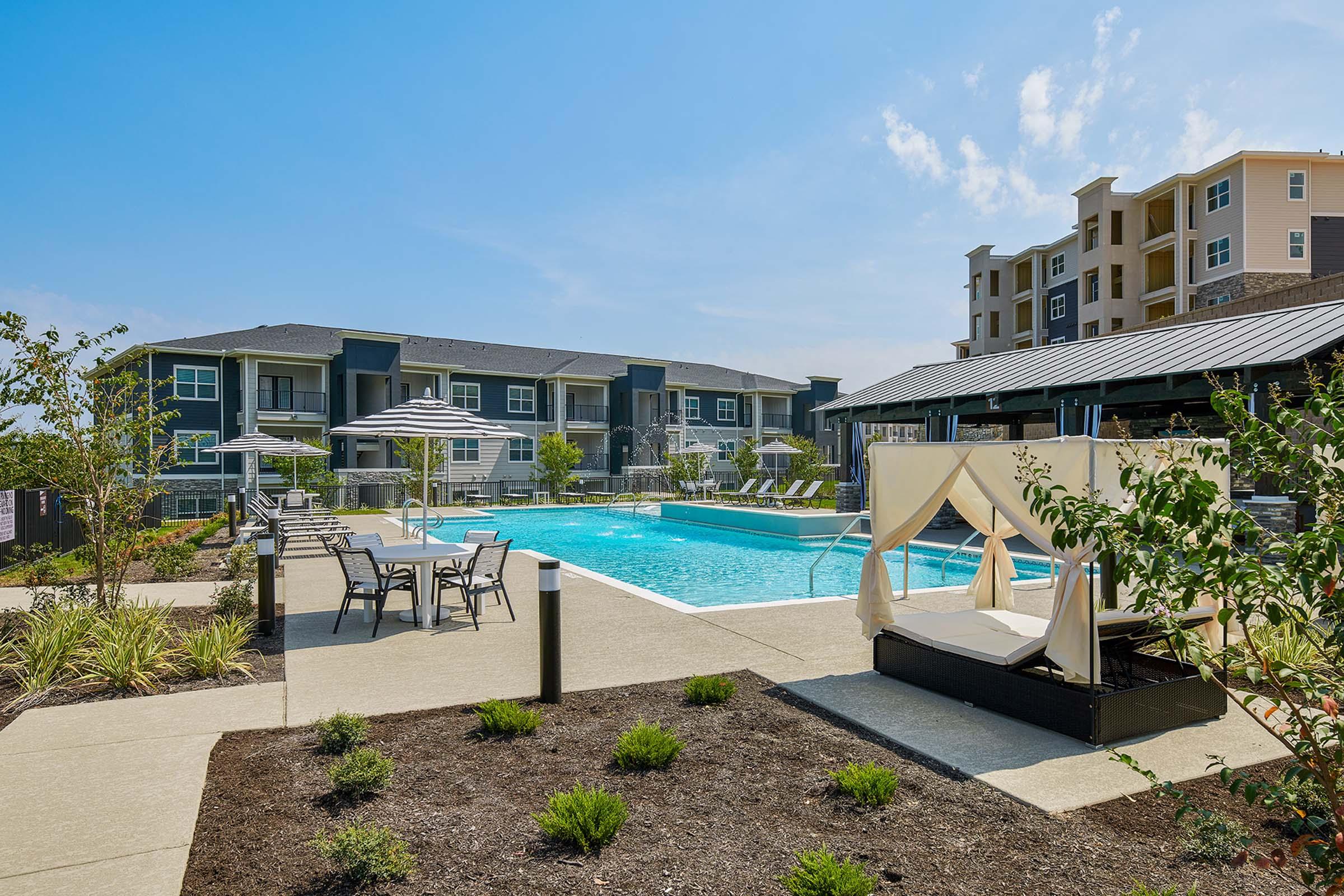 A landscaped outdoor pool area featuring a large swimming pool surrounded by lounge chairs and shaded umbrellas. Nearby, there are cabana-style seating areas and well-maintained greenery. Modern apartment buildings rise in the background under a clear blue sky.