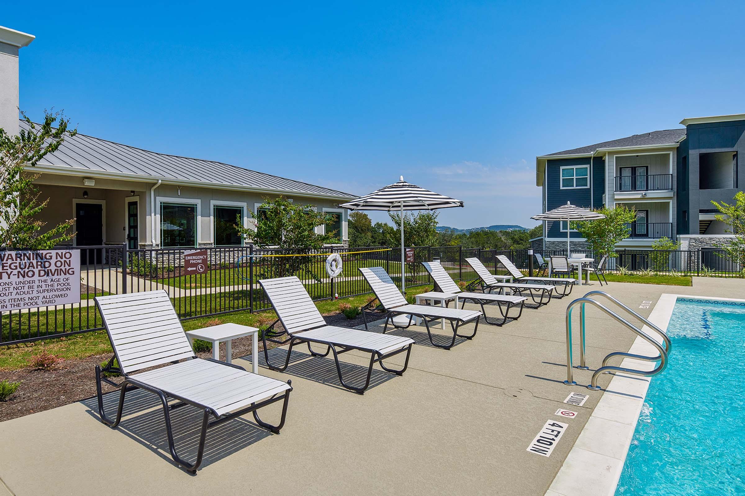 A scenic pool area featuring lounge chairs and umbrellas, surrounded by greenery. In the background, a residential building is visible under a clear blue sky. A sign indicating pool rules is posted nearby. The inviting pool is partially visible, showcasing a relaxing outdoor space.