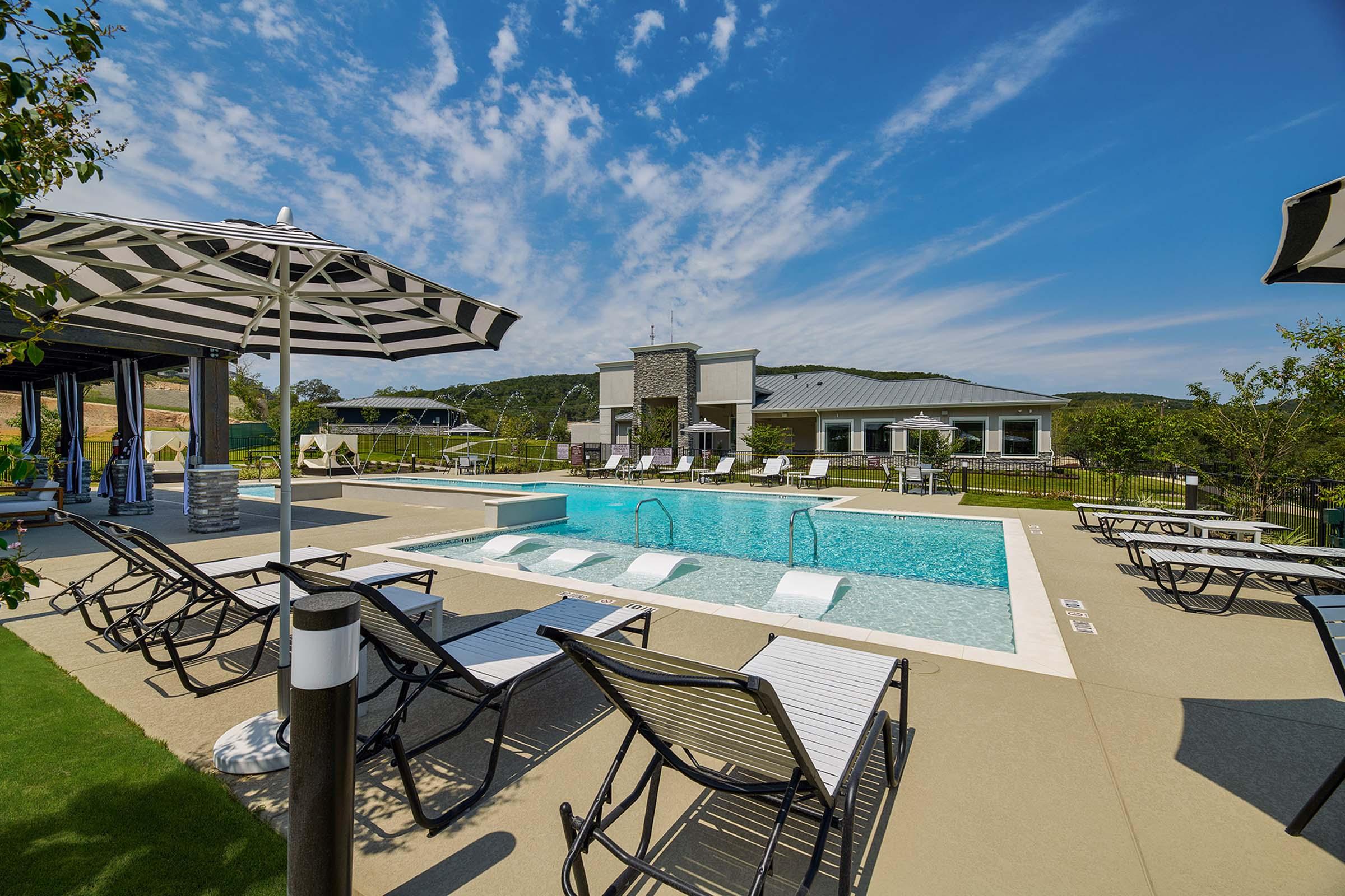 A bright, outdoor pool area featuring a clear blue pool with lounging chairs and striped umbrellas. In the background is a modern building with large windows, surrounded by lush greenery and rolling hills under a blue sky with scattered clouds.