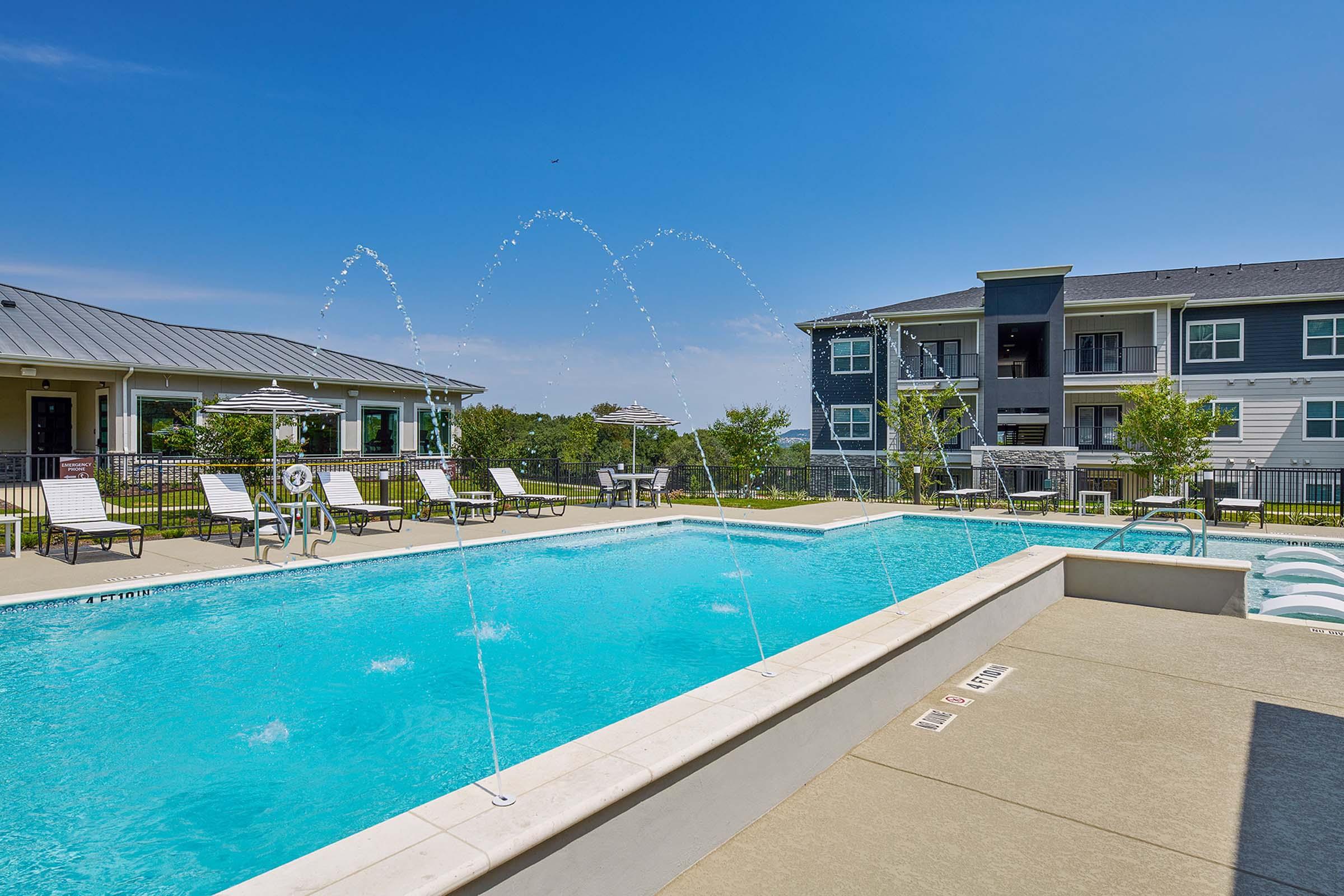 A clear blue swimming pool with water fountains is surrounded by lounge chairs and umbrellas. In the background, two multi-story residential buildings can be seen against a bright blue sky. Lush greenery adds to the serene atmosphere.