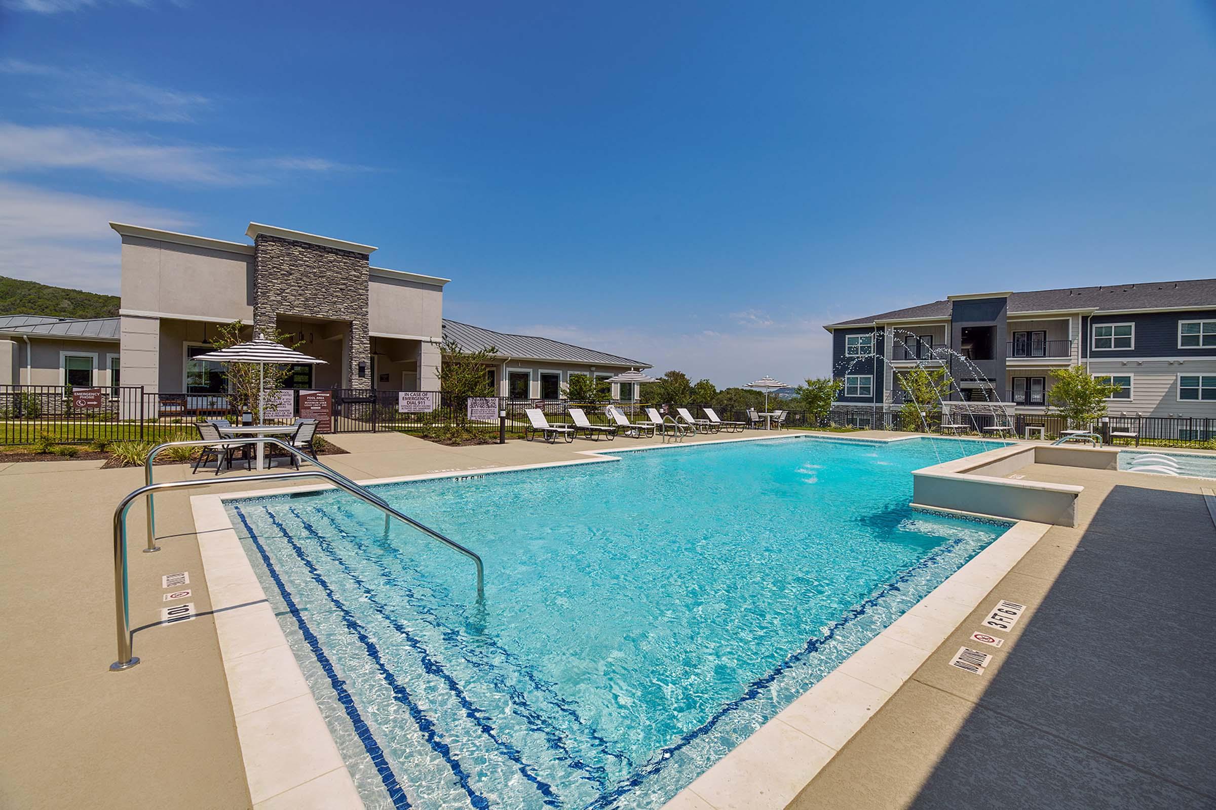 A clear blue swimming pool surrounded by lounge chairs and tables, with a modern building in the background. The area features lush greenery and a clear sky, creating a relaxing outdoor atmosphere perfect for leisure.