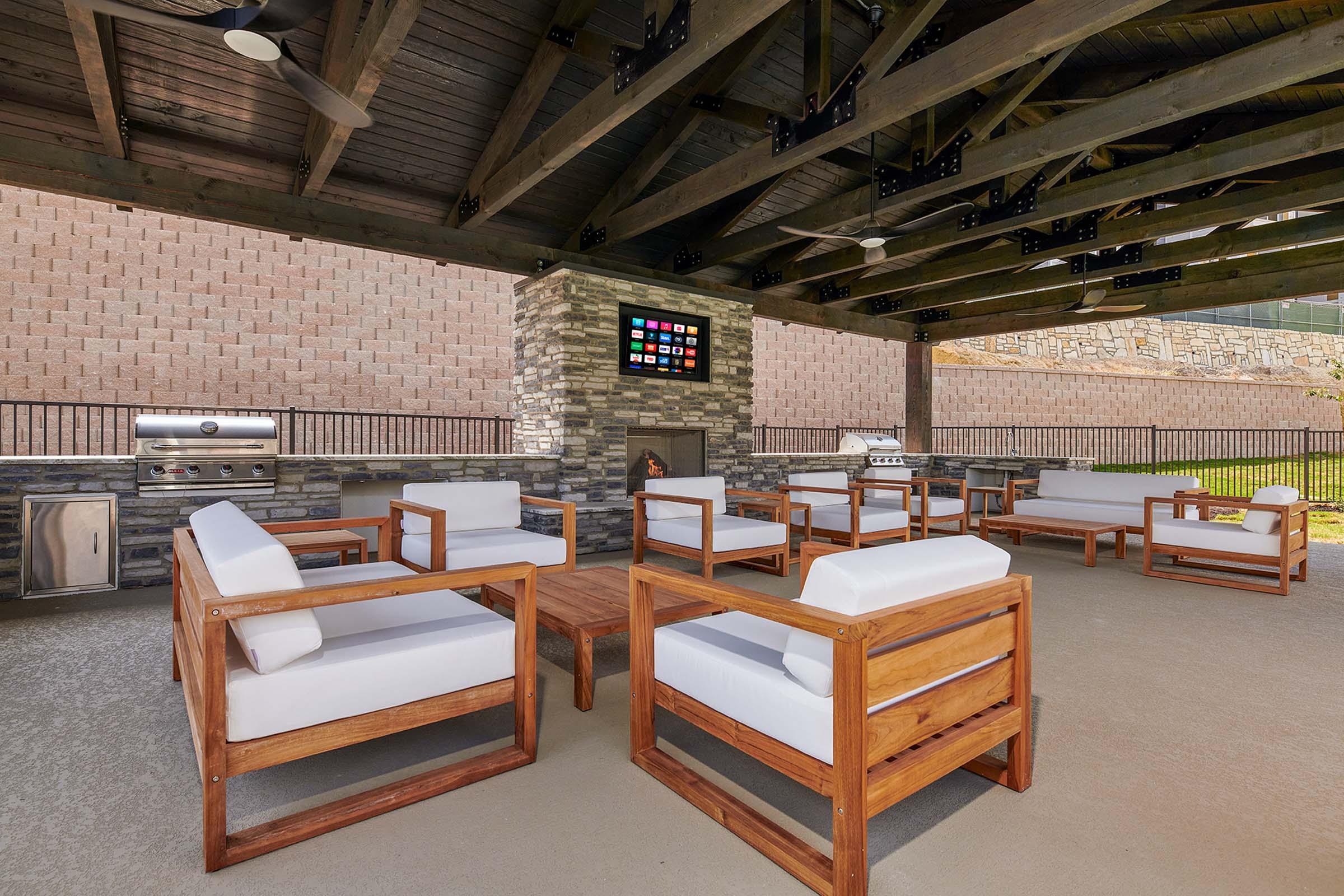 A cozy outdoor lounge area featuring wooden furniture with white cushions. There is a central coffee table, an outdoor grill station, and a stone fireplace. The space is shaded by a large wooden roof, and a flat-screen television is mounted on the wall, providing a comfortable setting for relaxation and entertainment.