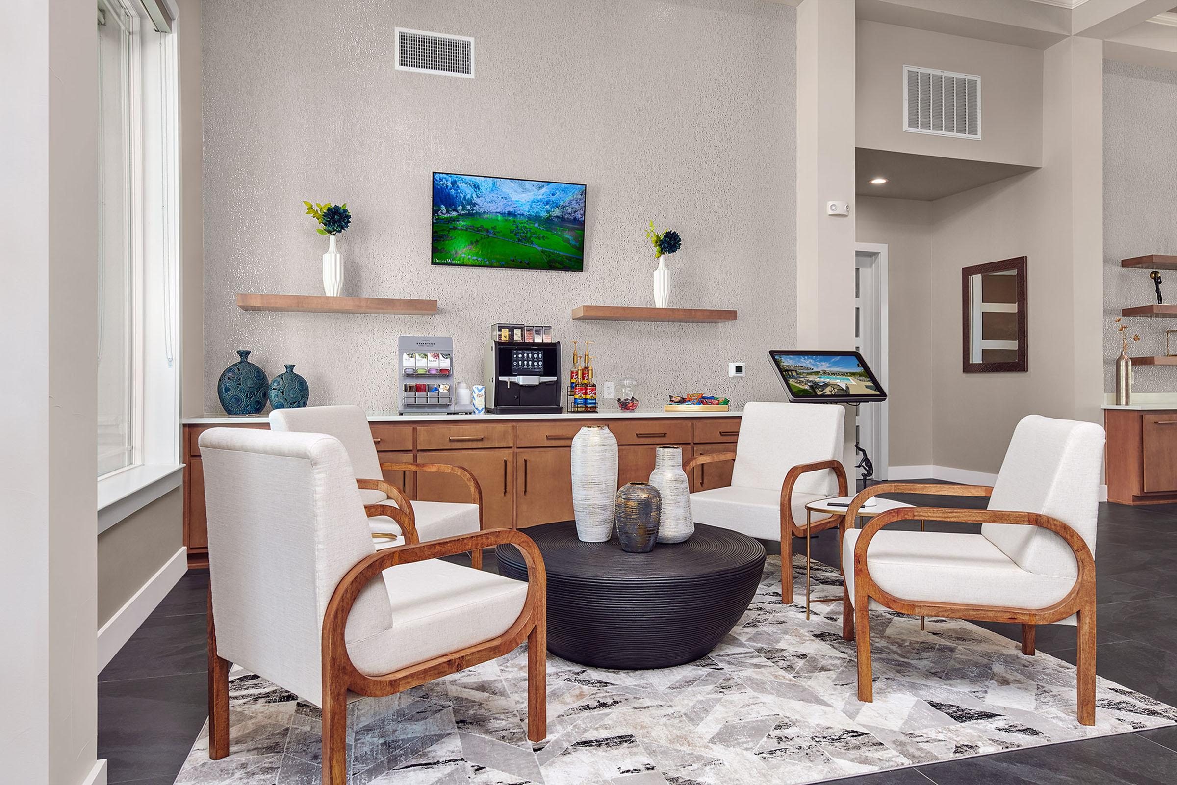 A modern lounge area featuring four comfortable white chairs with wooden frames surrounding a round black table. There is a patterned rug on the floor, a flat-screen TV mounted on the wall displaying a nature scene, and decorative plants and items on a side table and shelves.