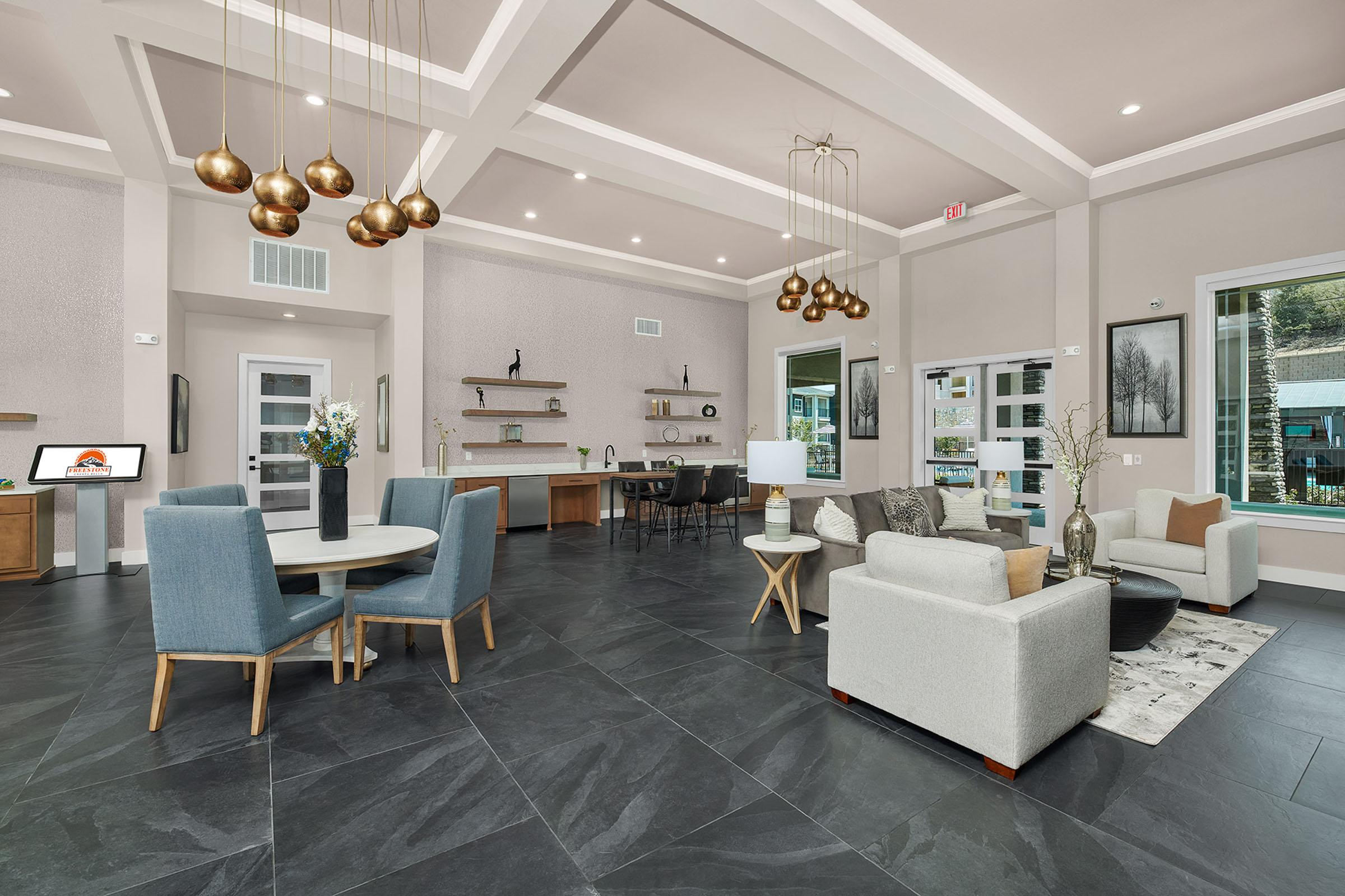 A spacious and modern lobby featuring a round dining table with four chairs, contemporary sofas, and decorative lighting. The floor is dark slate, and large windows allow natural light to flood the room, which includes wooden shelves and stylish decor elements, creating a welcoming atmosphere.