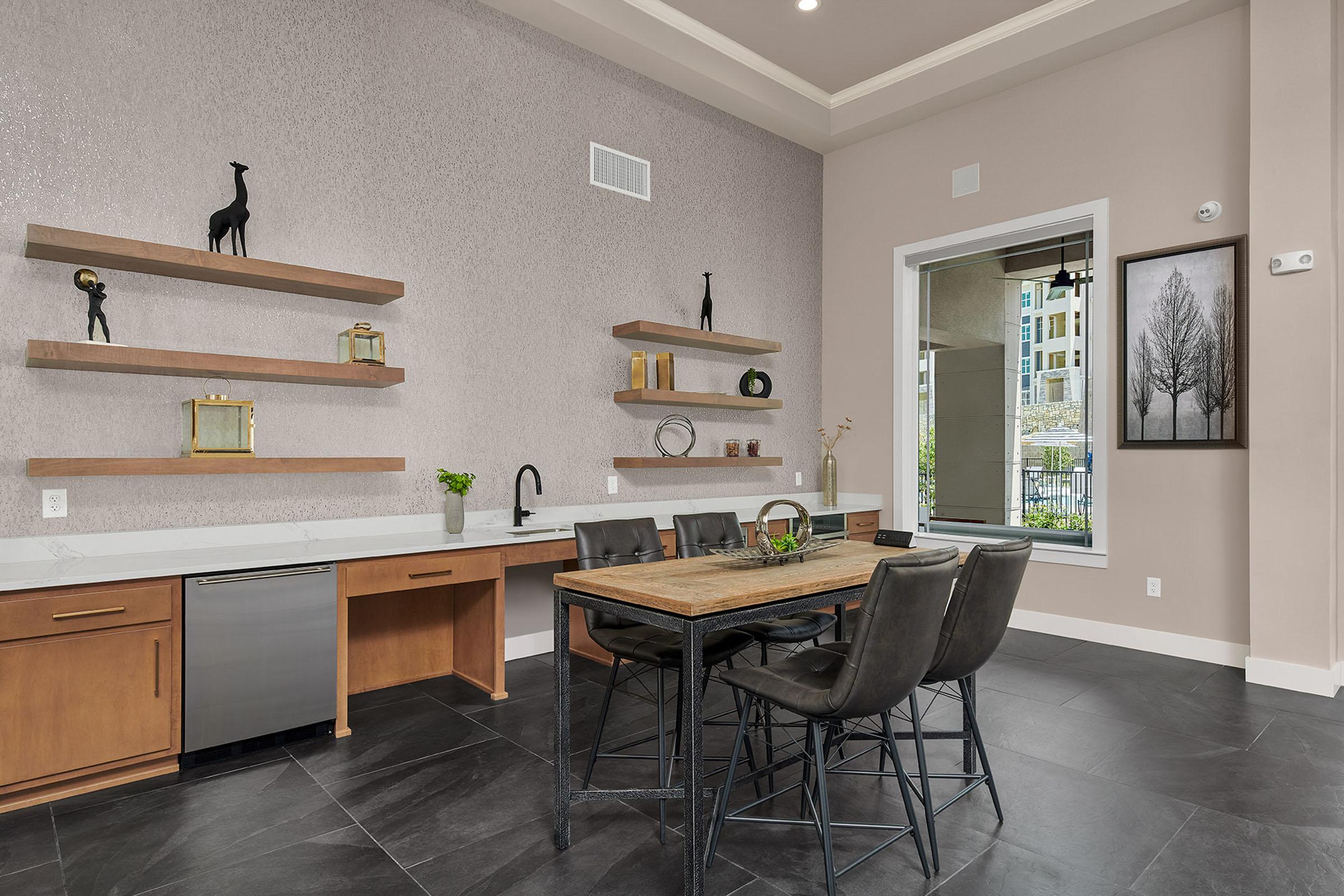 Modern dining area featuring a wooden table surrounded by black chairs. Wall-mounted shelves with decorative items and plants are displayed above a sleek kitchen area with a stainless steel refrigerator. Large windows allow natural light to fill the space, showcasing a view of the outdoors.