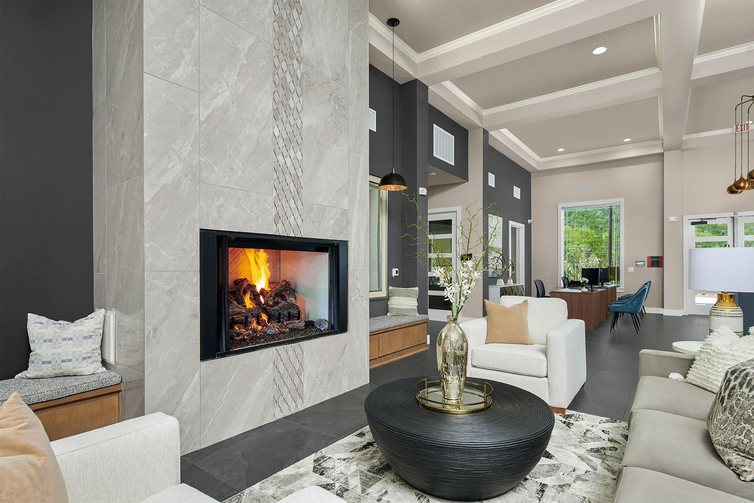 A modern living room featuring a sleek fireplace with a flames, stylish furniture including a light-colored sofa, a round table with a black base, and decorative accents. The space has a contemporary design with neutral colors, hardwood floors, and large windows allowing natural light.