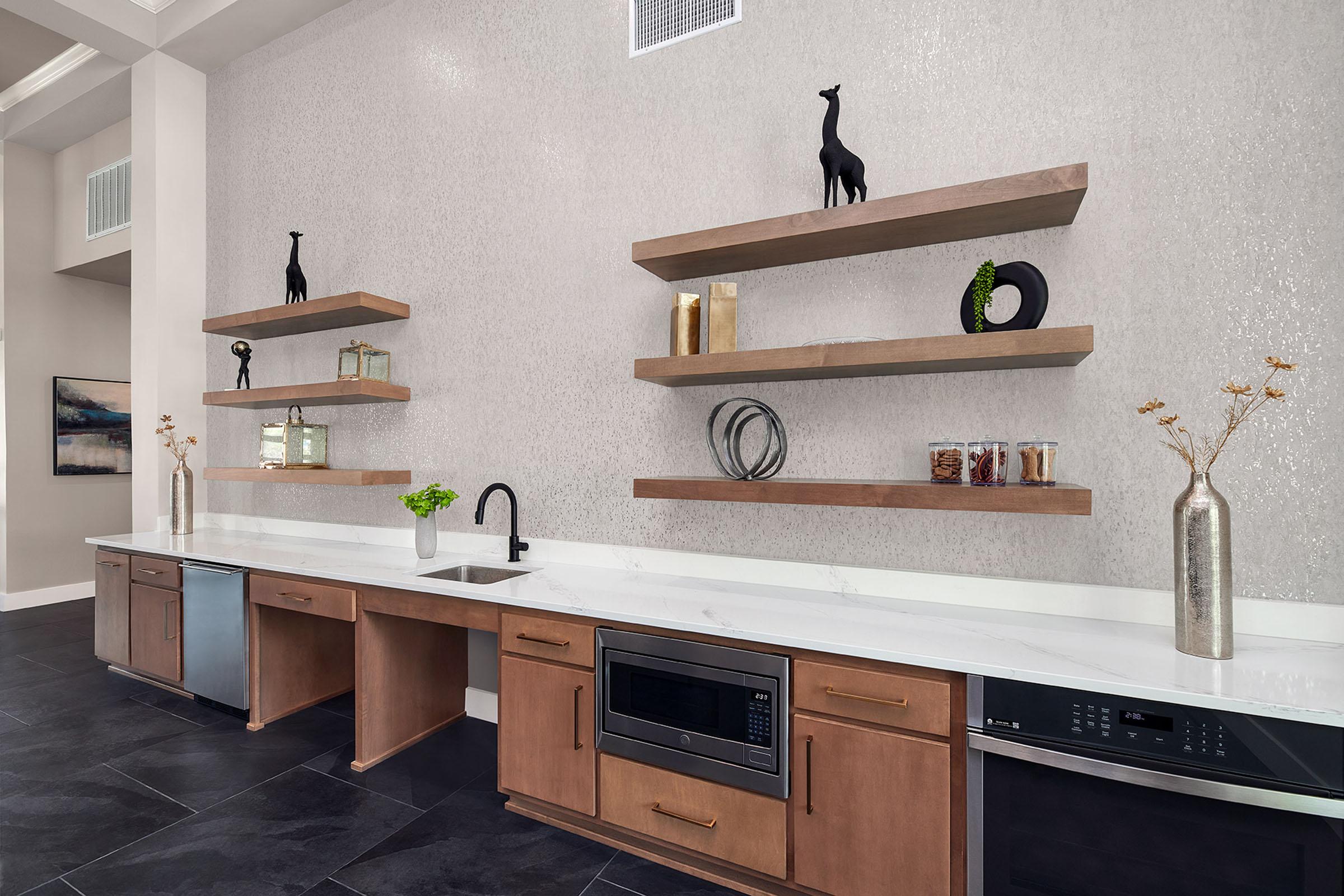 A modern kitchen with dark tile flooring and light-colored walls. The countertop features a sleek sink, a microwave, and a built-in dishwasher. Above, stylish wooden shelves hold decorative items, including small giraffe figurines, glass containers, and a vase with flowers, adding a contemporary touch to the space.