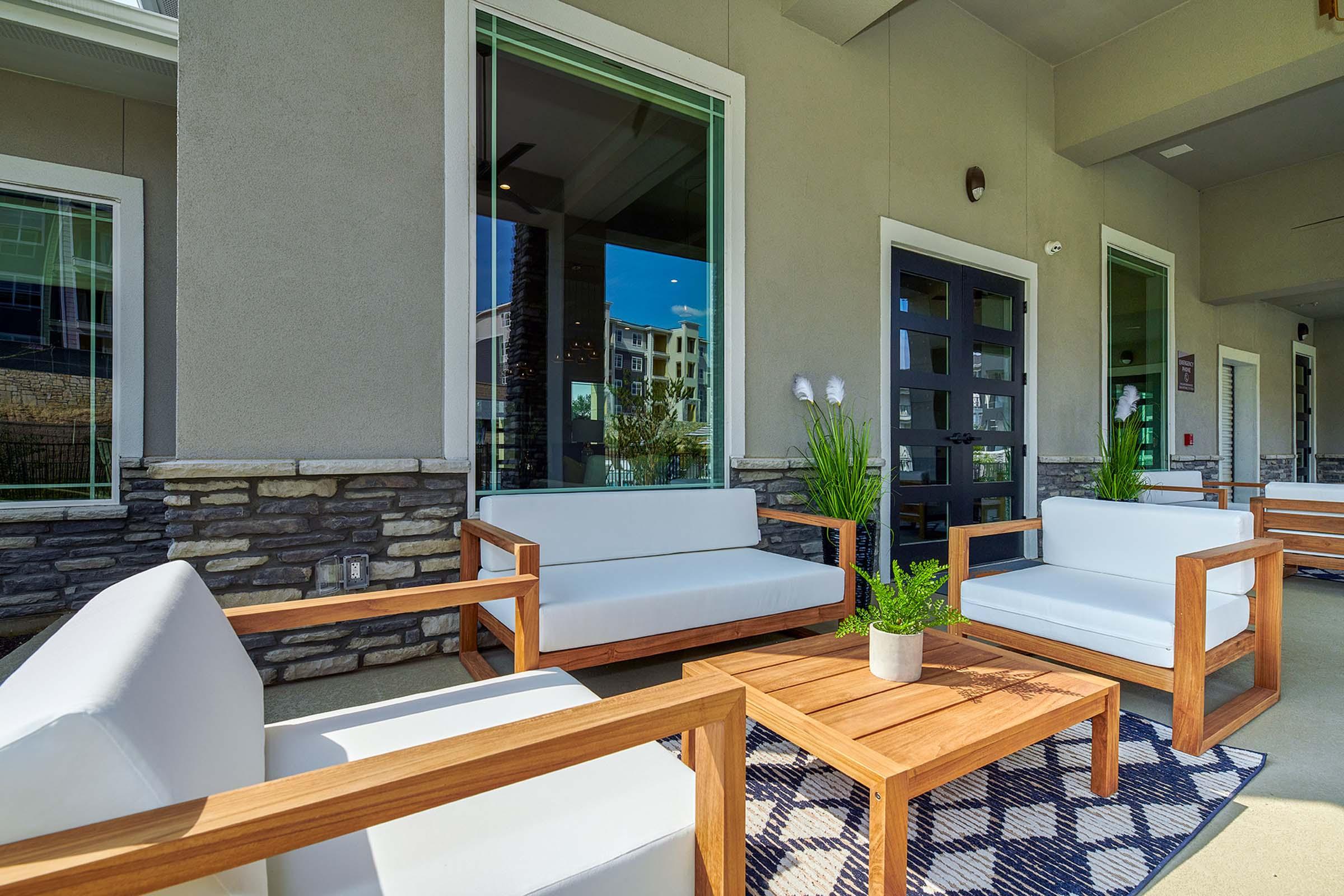 A modern outdoor seating area featuring white cushioned chairs and a wooden coffee table with a small potted plant. The space has large windows showcasing an exterior view, with a stone accent wall adding texture to the overall design. Sunlight illuminates the area, creating a welcoming ambiance.