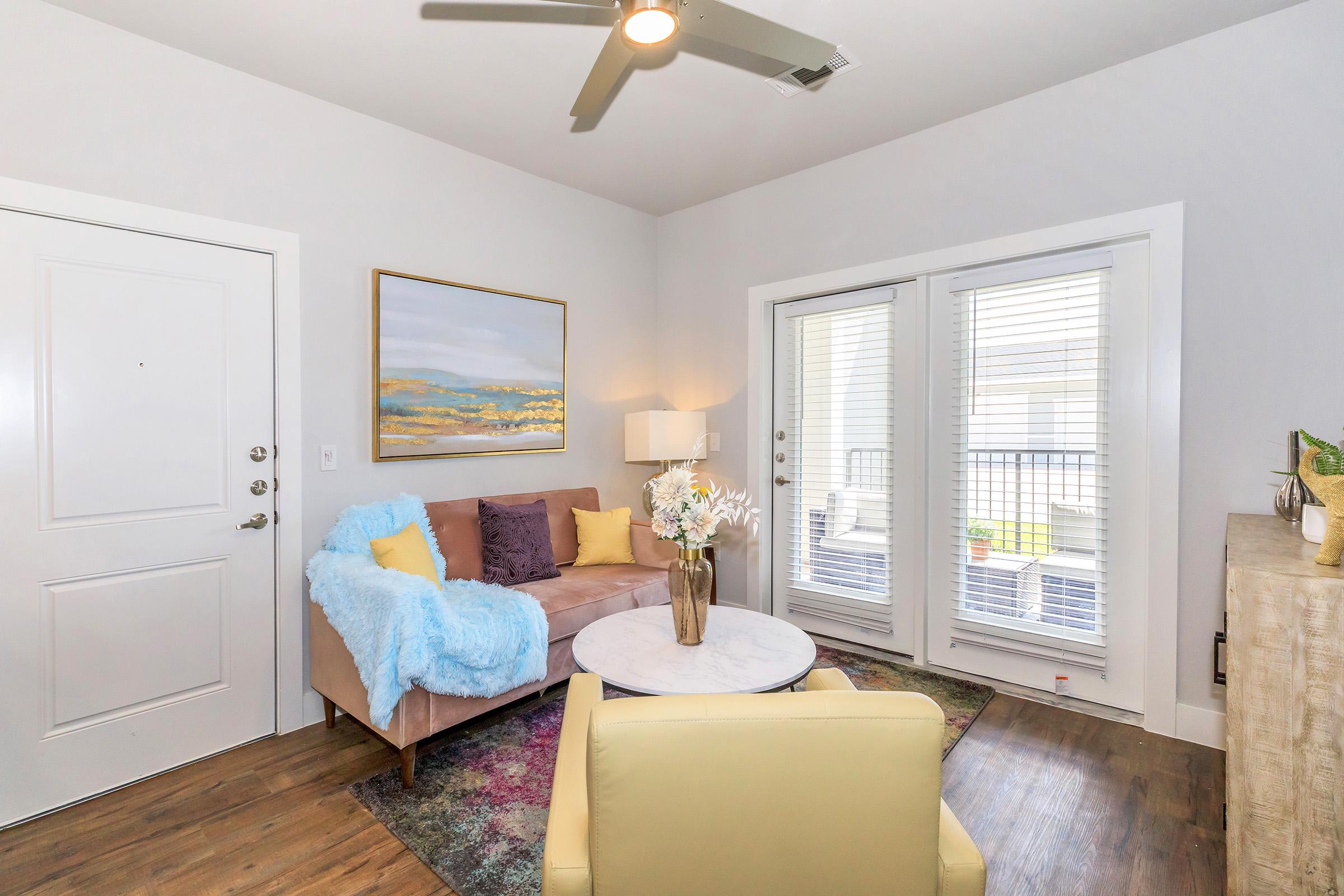 Cozy living room featuring a light-colored sofa with decorative pillows, a round coffee table, and a lamp. Large windows with blinds allow natural light, and an artwork hangs on the wall. A small porch is visible through the sliding glass doors, adding to the inviting atmosphere of the space.