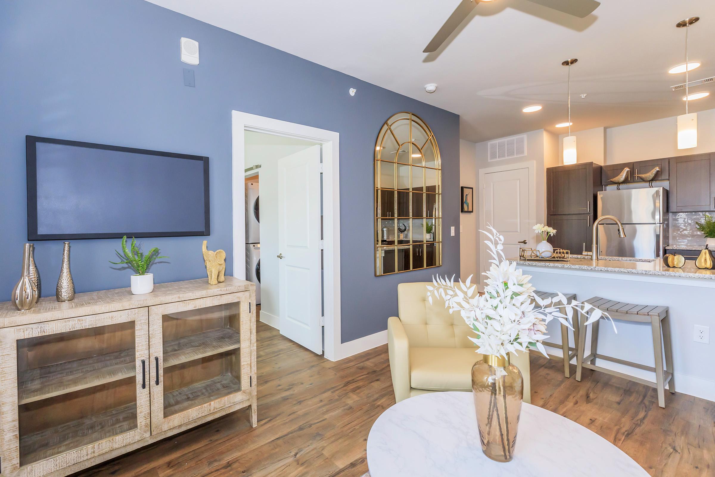 A stylish living room featuring a light-colored coffee table with a floral arrangement, a beige armchair, and a wooden cabinet. The walls are painted dark blue, and there is a large gold-framed mirror. A kitchen area is visible with modern appliances, and a doorway leads to another room. Bright lighting enhances the space.