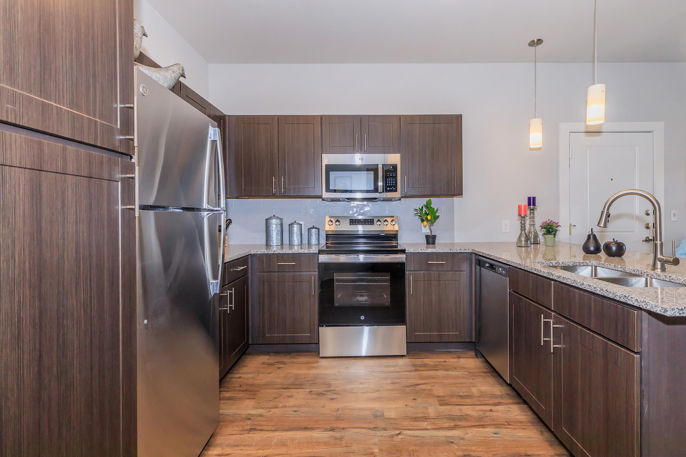 Modern kitchen featuring dark wood cabinetry, stainless steel appliances including a refrigerator, microwave, and oven. Granite countertops complement the sleek design. The space is well-lit with pendant lighting and includes decorative elements such as a vase and candles, creating a warm and inviting atmosphere.