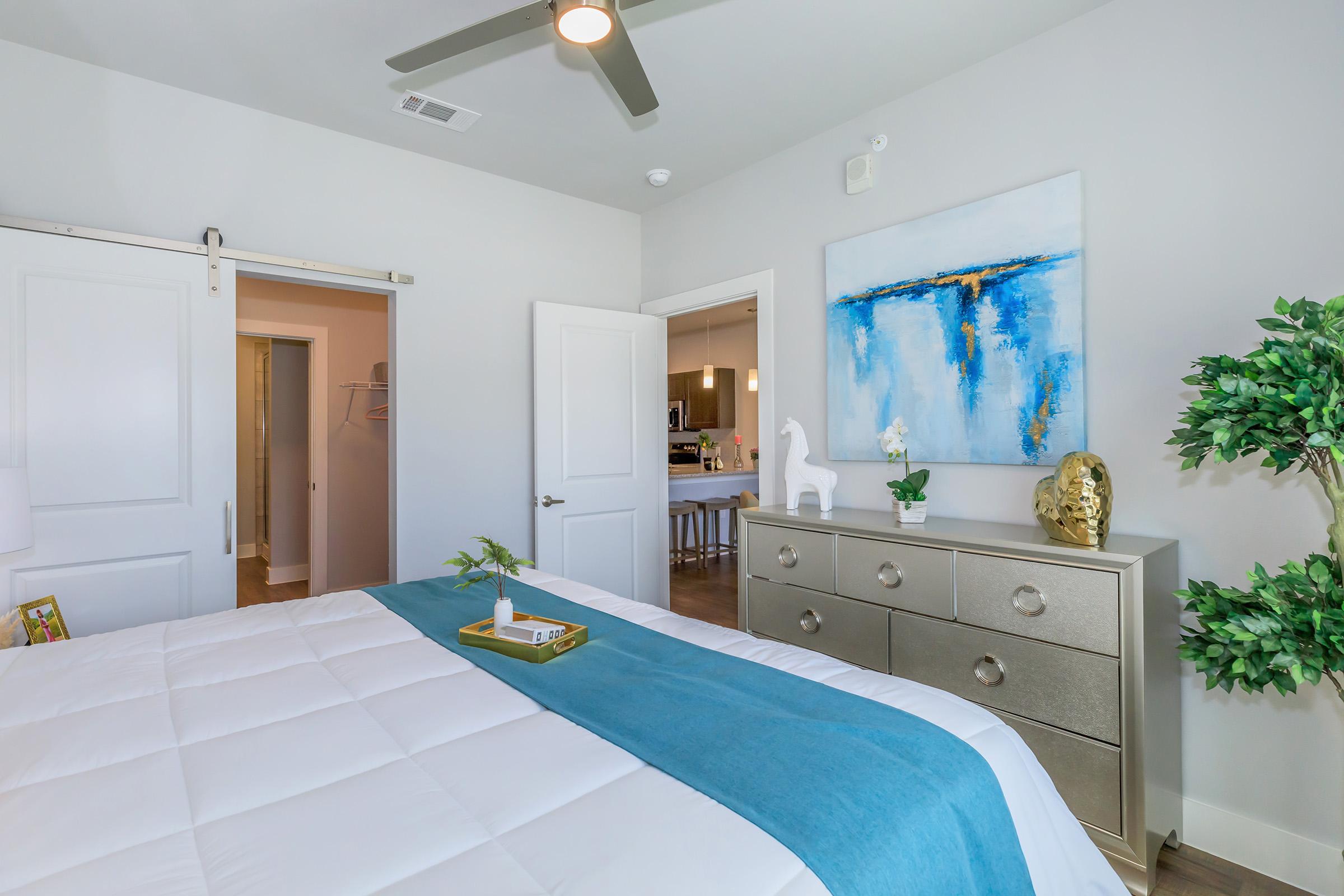 A modern bedroom featuring a king-sized bed with a light blue blanket, a decorative tray on the bed, a stylish dresser with a golden sculpture, and a plant. There are white walls and a sliding door leading to another room. Bright natural light floods the space, creating a fresh and inviting atmosphere.