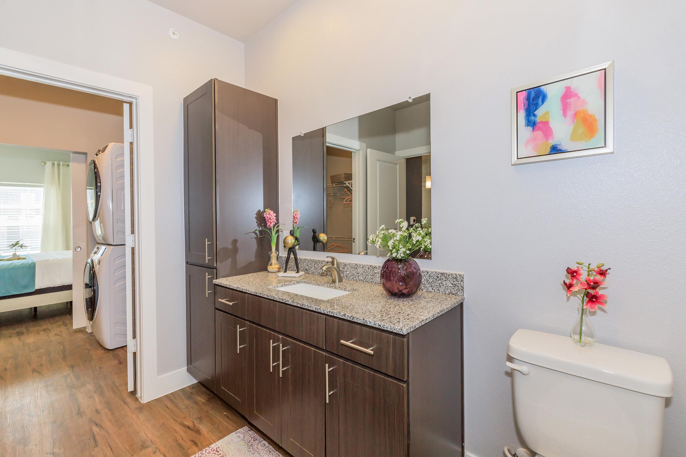 A modern bathroom featuring a dark wood vanity with a granite countertop, a large mirror above the sink, and decorative flowers. A toilet is visible to the right, and a doorway leads to another room in the background. The walls are painted in a light gray color, and the floor is wooden.