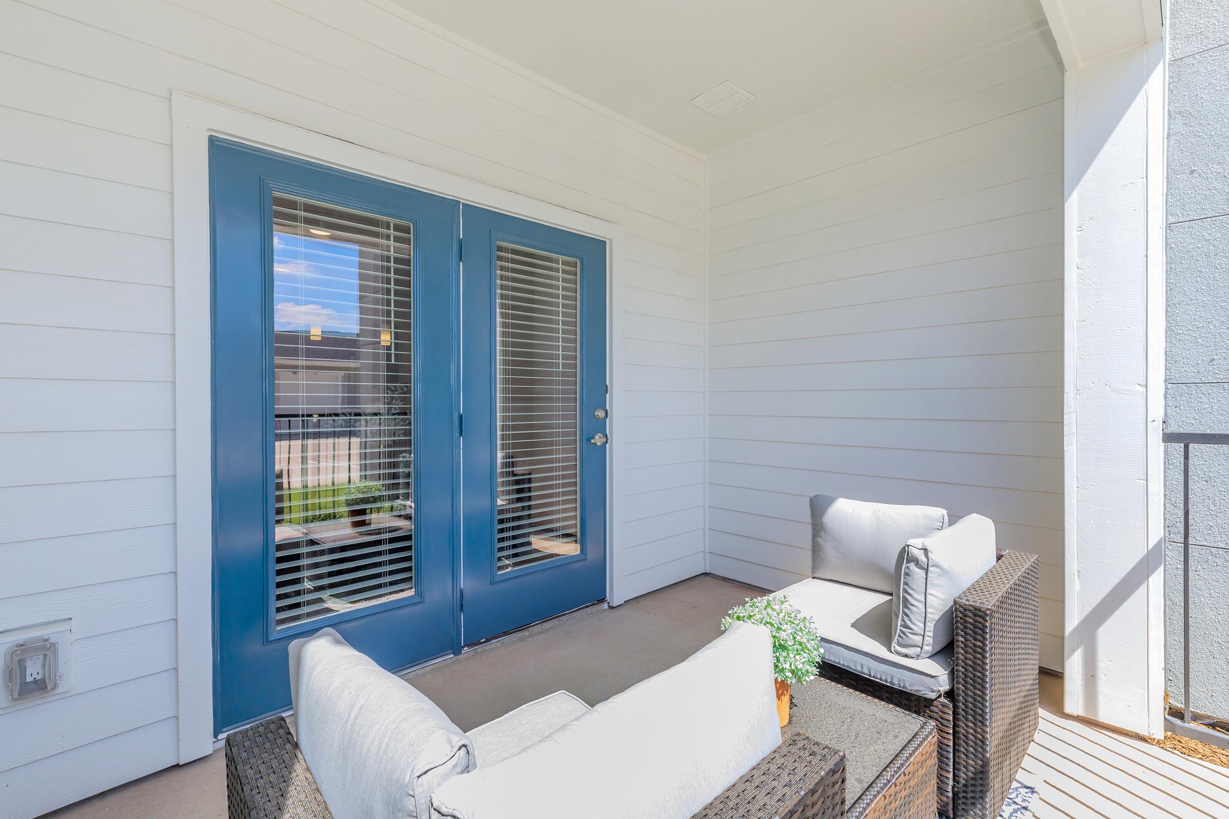 A small outdoor balcony or patio featuring two gray cushioned chairs and a matching table, with a decorative plant. There are double blue doors with vertical slats leading inside, set against a white-paneled wall, and bright sunlight illuminates the space.
