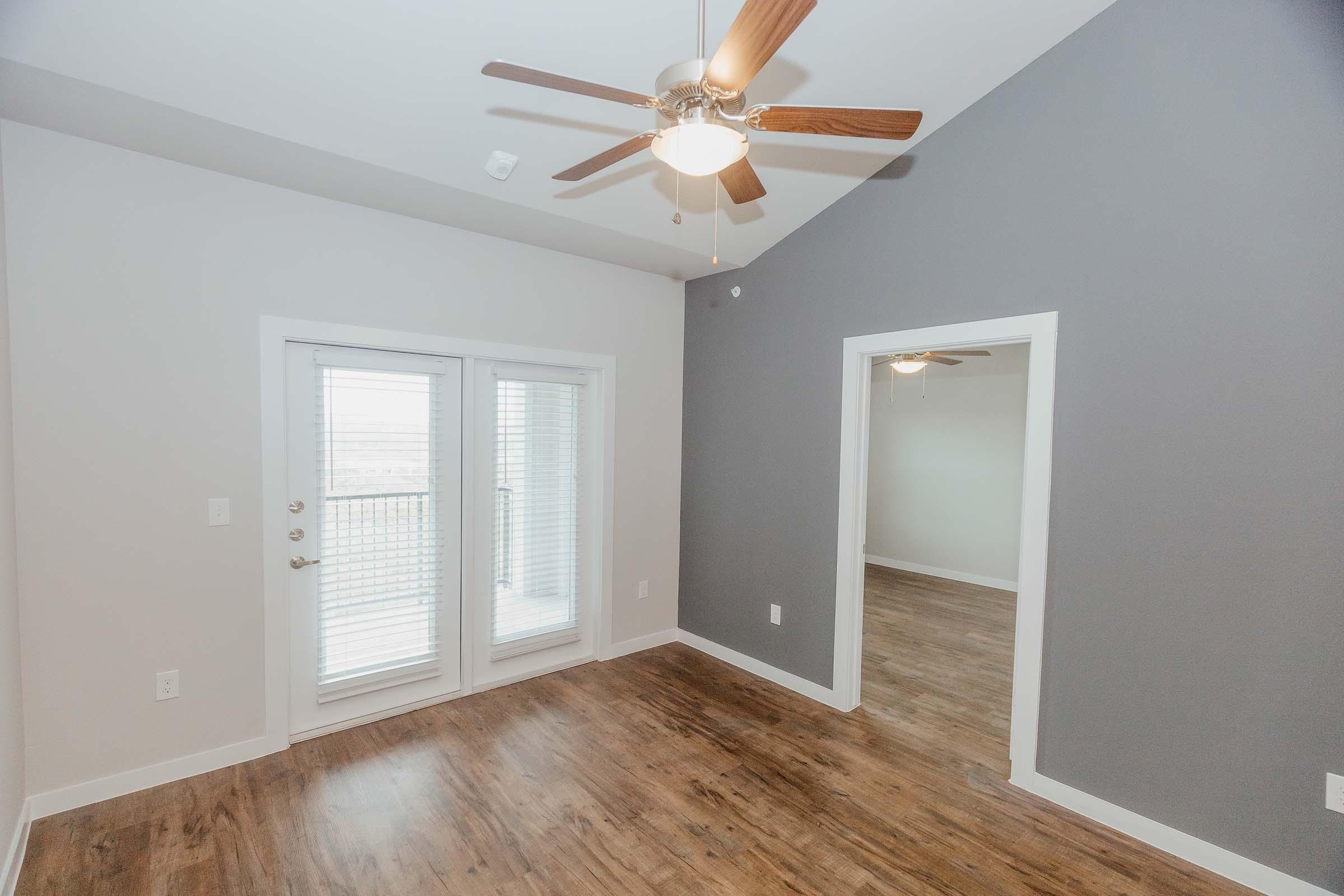 Interior view of a room featuring light gray walls and a ceiling fan with wooden blades. There are double doors leading to a balcony, and a hardwood floor adds warmth to the space. A second room is visible in the background, with a similar ceiling height and design.