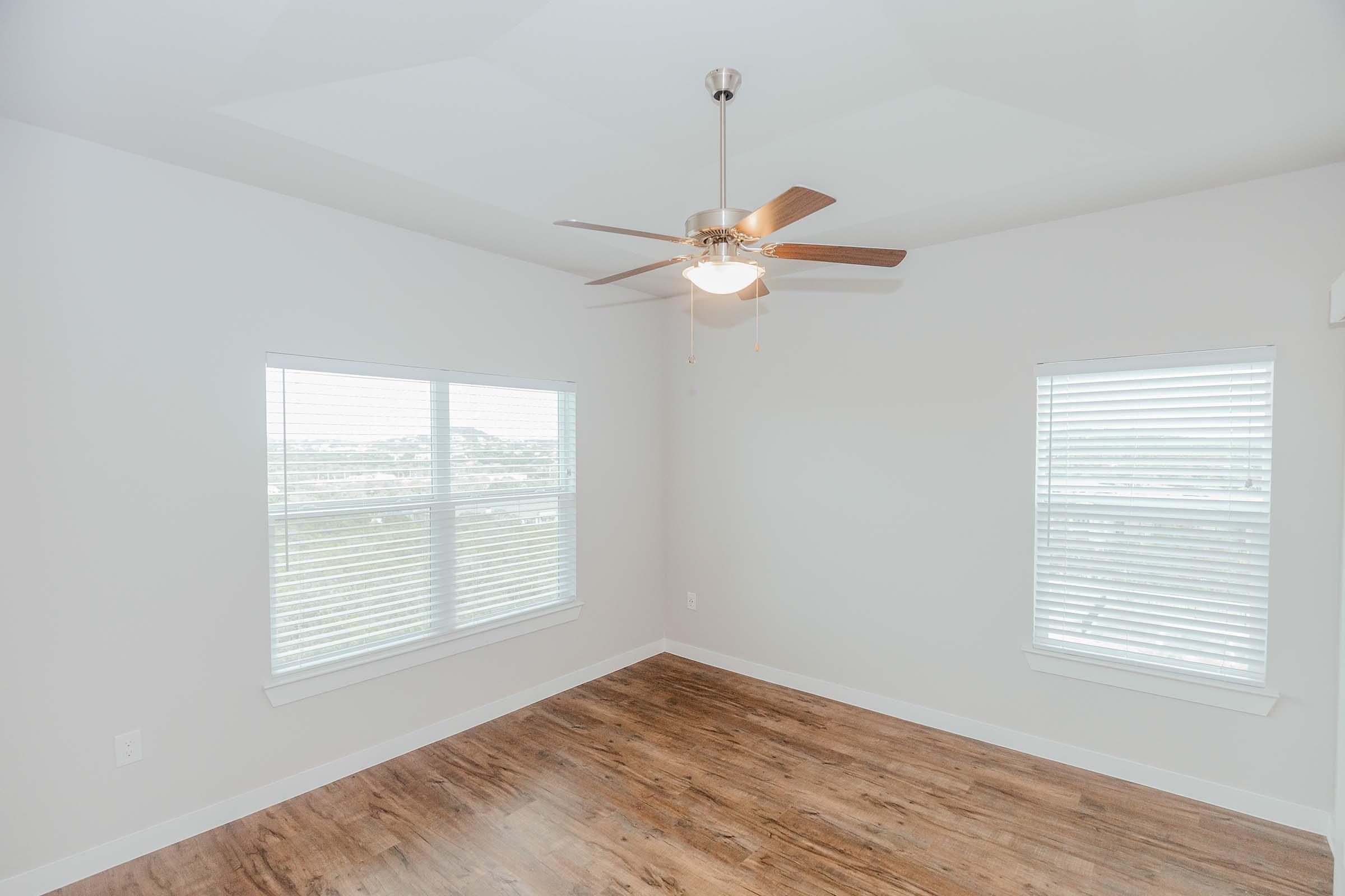 A bright, empty room featuring light-colored walls and a wooden floor. There are two windows with white blinds allowing natural light to enter. A ceiling fan with wooden blades is mounted in the center of the room, creating a spacious and airy feeling.