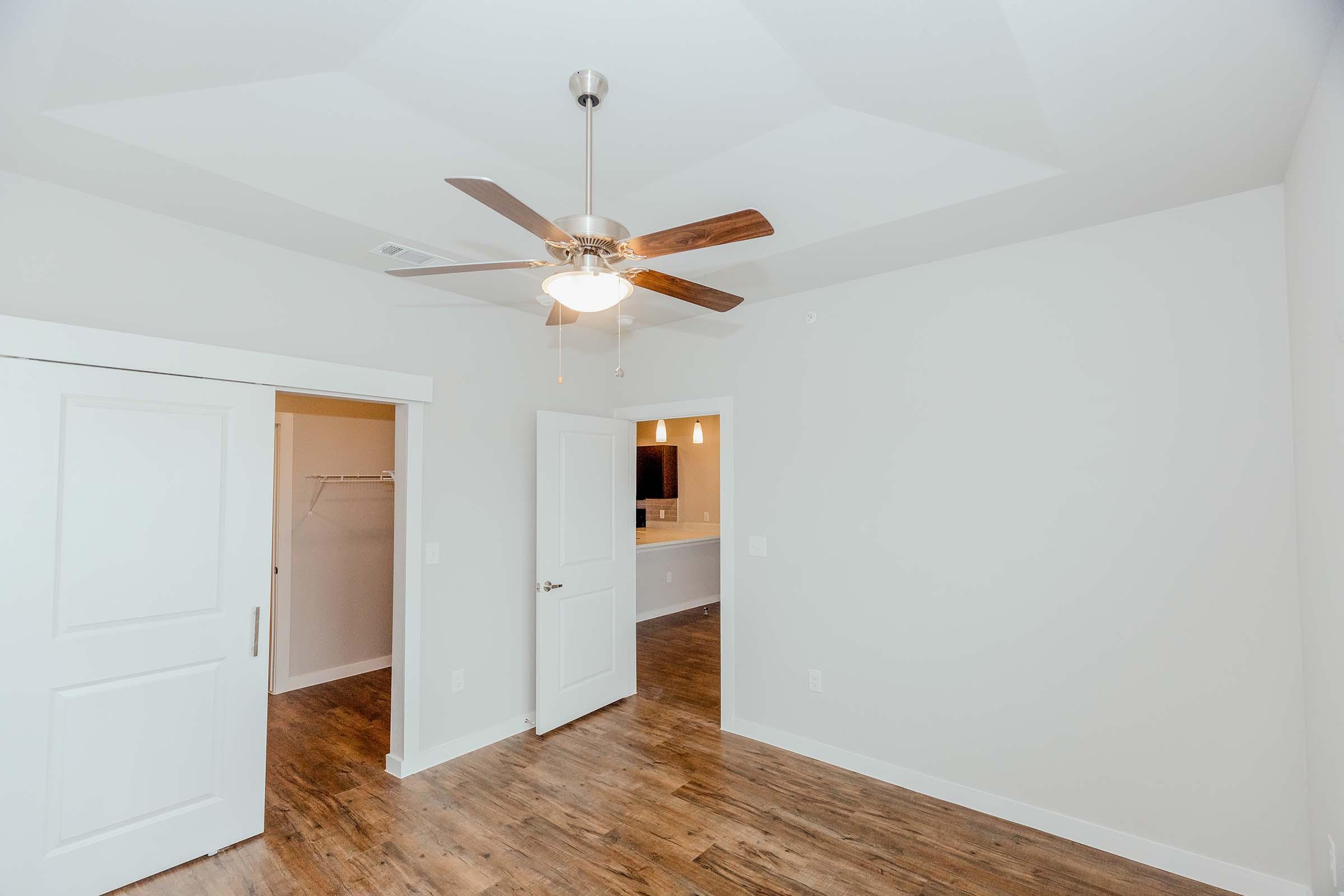 A well-lit empty room featuring a ceiling fan with wooden blades, light-colored walls, and a hardwood floor. Two doors are visible: one leading to a closet and another to another area. The background shows a glimpse of a kitchen with light fixtures. The space is modern and inviting.