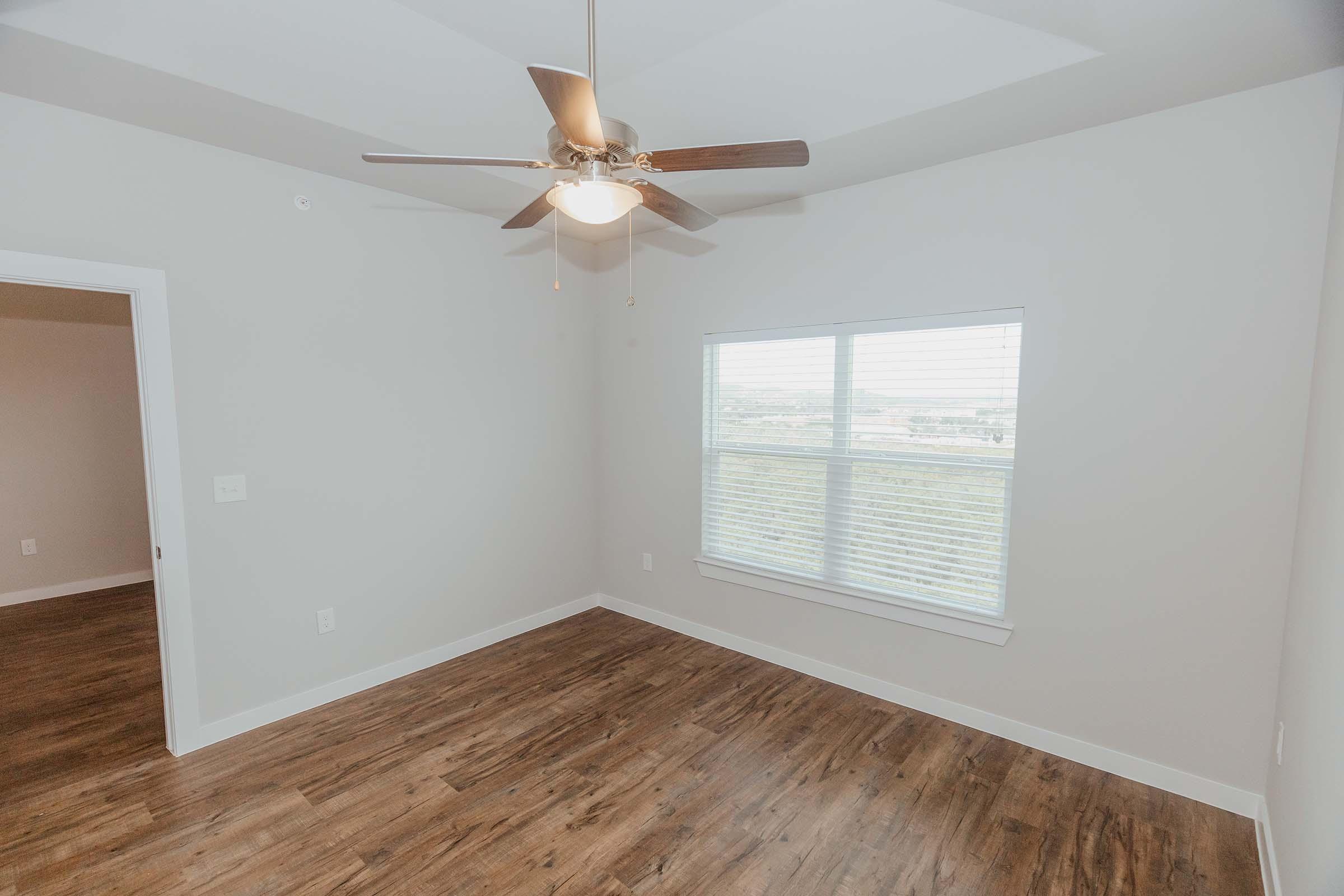 A well-lit, empty room with light-colored walls and wood-style flooring. A ceiling fan is installed in the center, and there is a window on one wall allowing natural light to enter. The room appears spacious and is ready for furnishing.