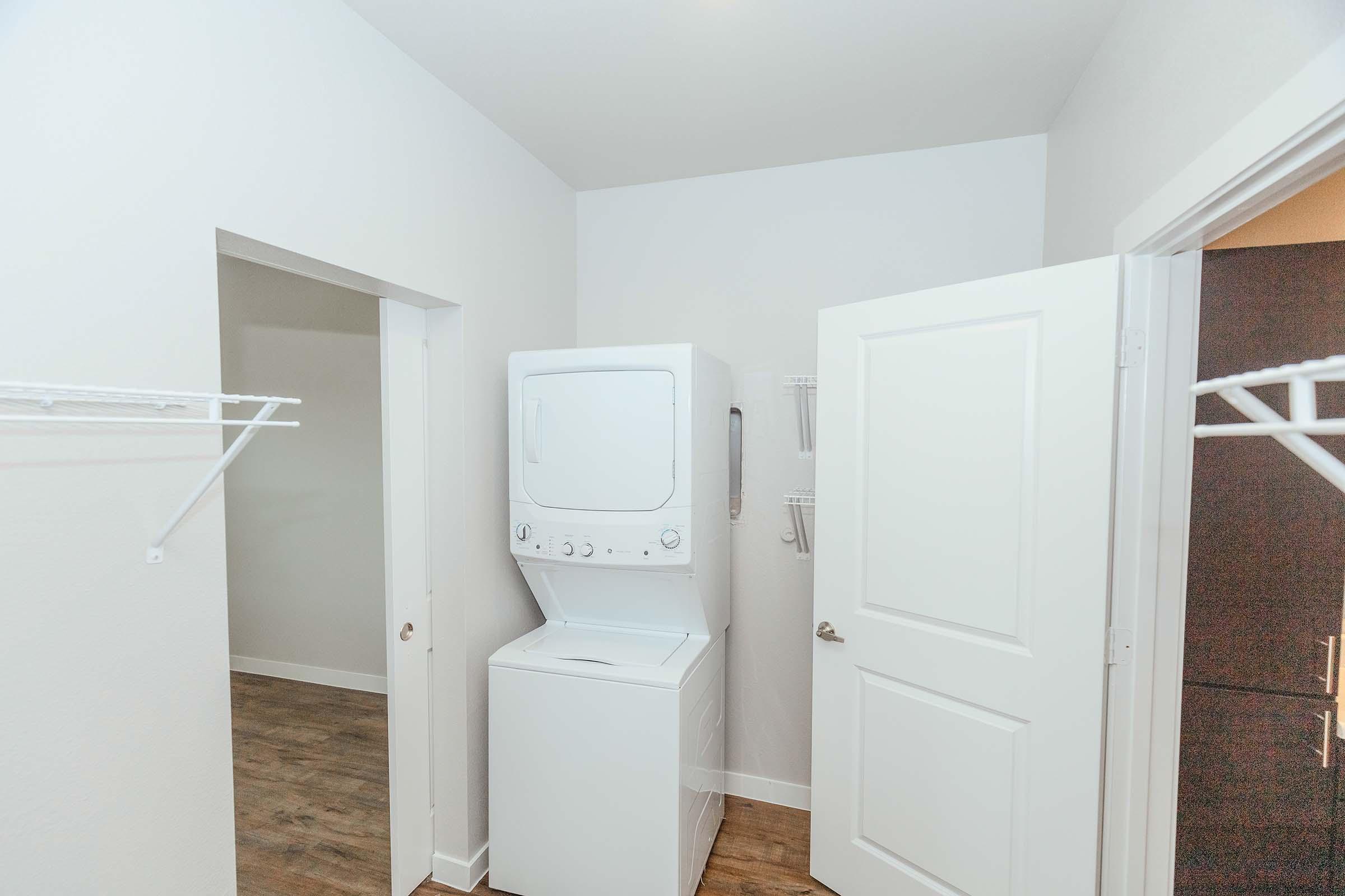 A laundry room featuring a stacked washer and dryer unit against a light-colored wall. Two open doors lead to adjacent spaces, and there are white wire shelves on either side for storage. The floor is covered in light wood laminate.