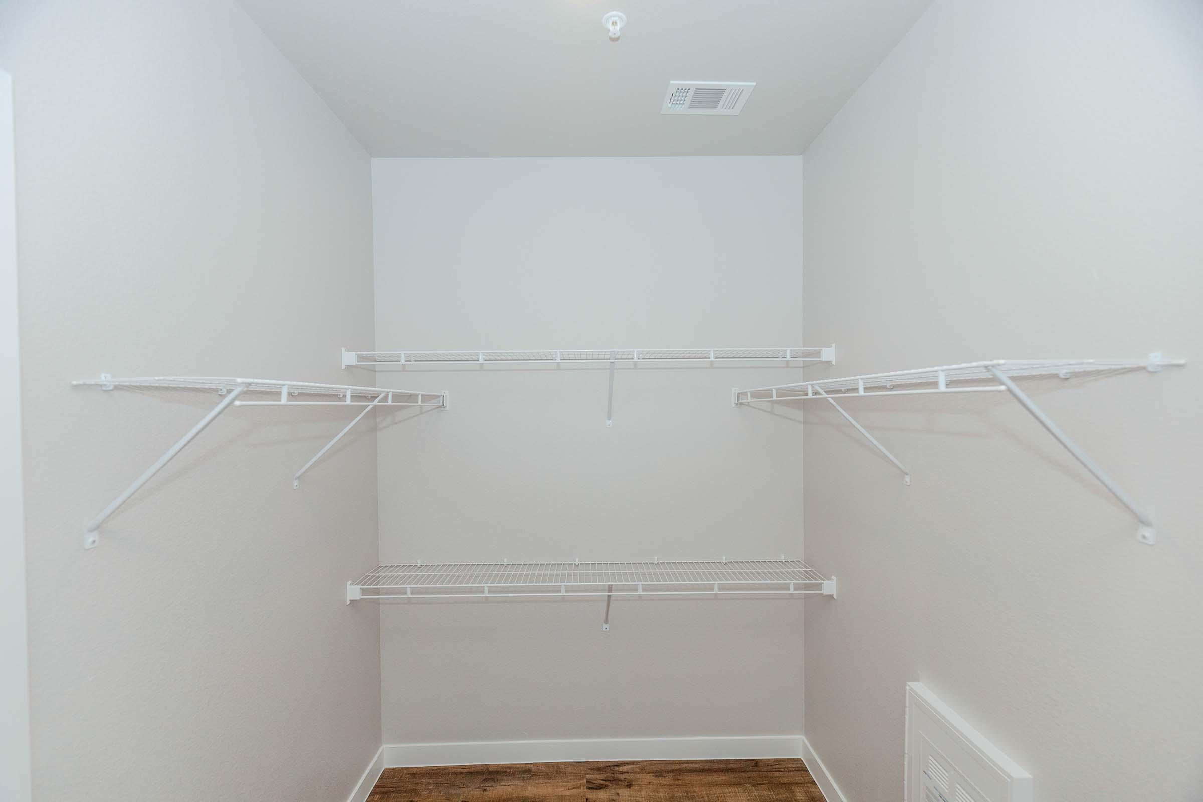 Empty closet with white wire shelving mounted on light gray walls. The floor is a warm wood tone, and there is an air vent on the lower right side. The overall appearance is clean and minimalistic, with ample space for storage.