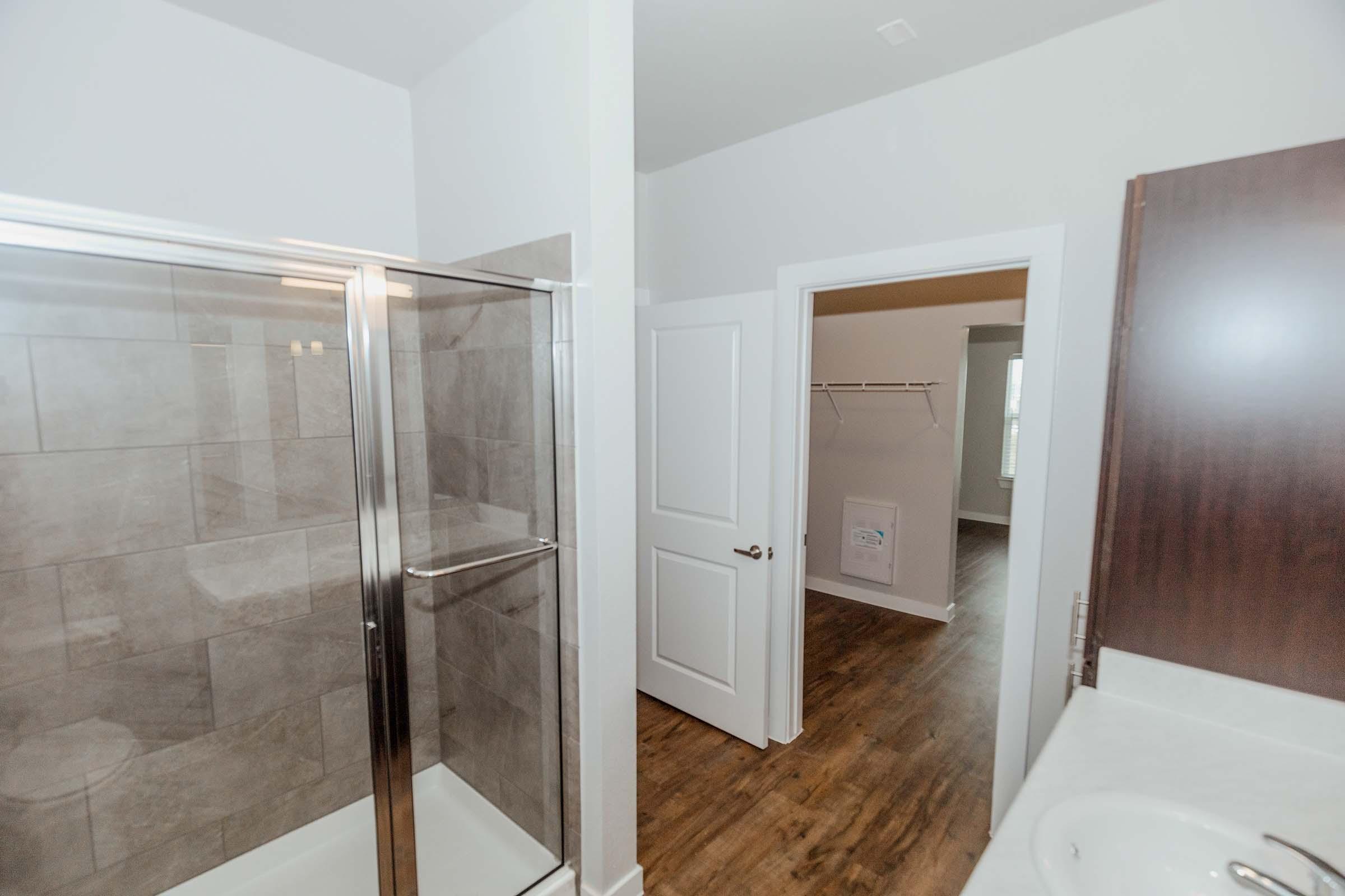 A modern bathroom featuring a glass shower enclosure with tile walls, adjacent to a doorway leading to a spacious room with a carpeted floor and a closet. The bathroom includes a counter with a sink and minimalistic cabinetry, set against light-colored walls and warm wood flooring.