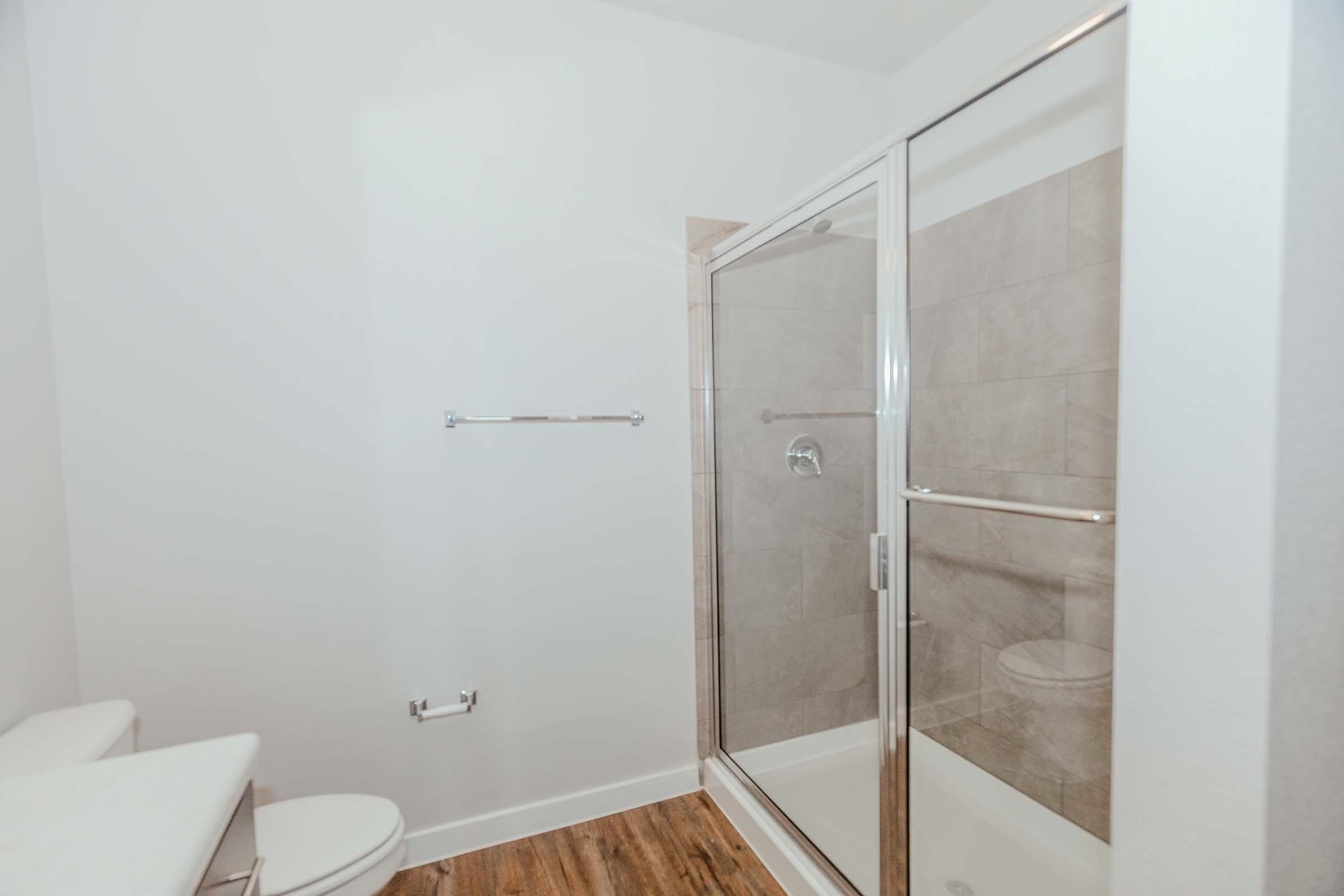 Modern bathroom featuring a glass shower enclosure, a single toilet, and a countertop with a sink. The walls are light-colored, and the floor has wood-like tiles. A towel bar is mounted on the wall, and the shower area has tiled walls. The overall design is clean and minimalist.