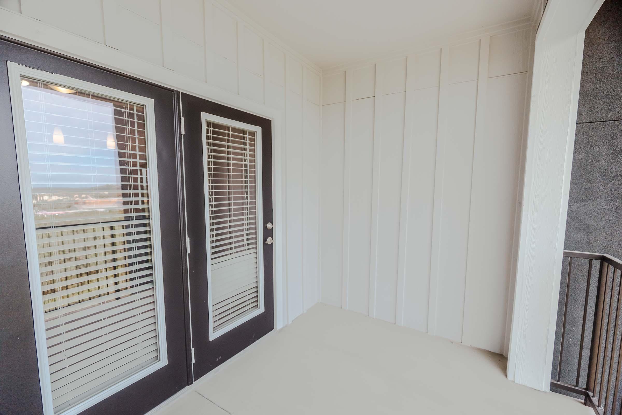 An empty balcony featuring sliding glass doors with blinds, light-colored walls, and a neutral floor. There is a railing visible on one side, showing a view beyond the balcony, suggesting a modern outdoor space in a residential building.