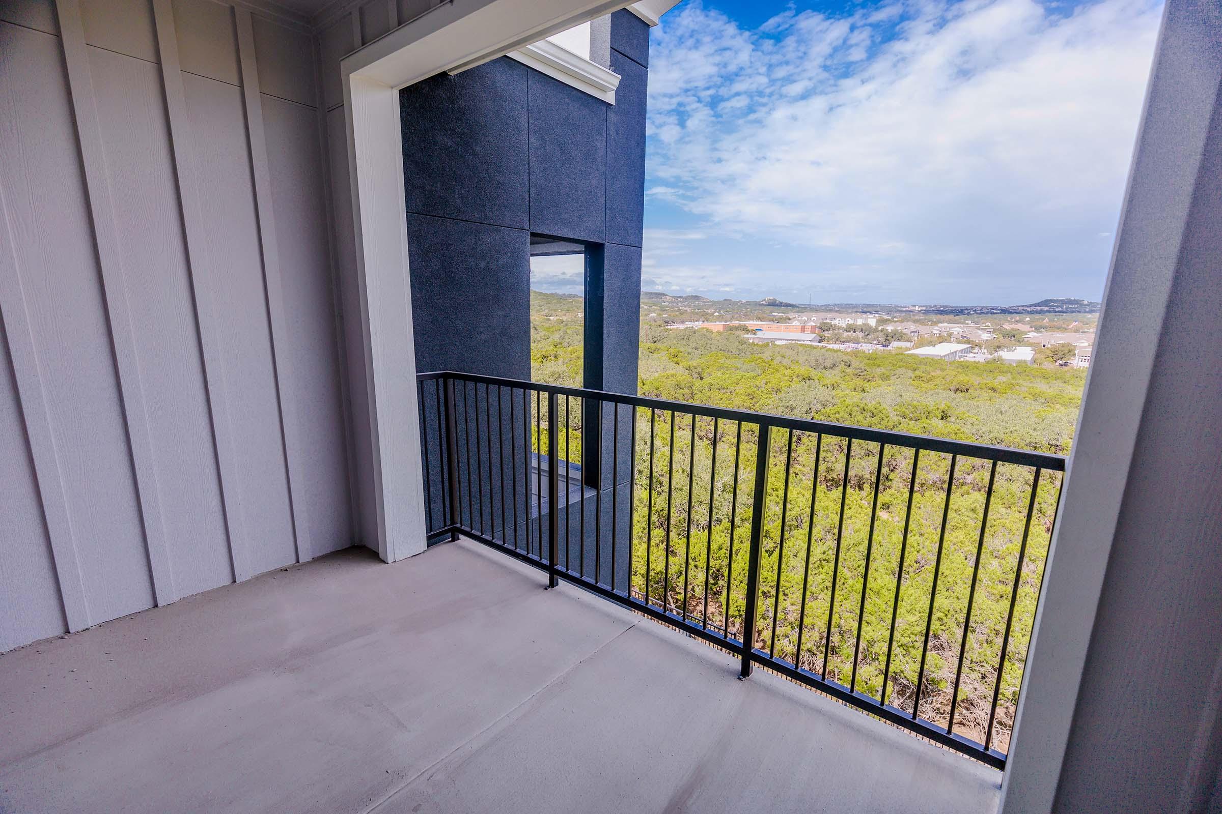 A modern balcony with a black railing, overlooking a scenic view of green hills and blue sky. The setting suggests a bright, open outdoor space surrounded by nature, with a hint of residential structures in the distance.