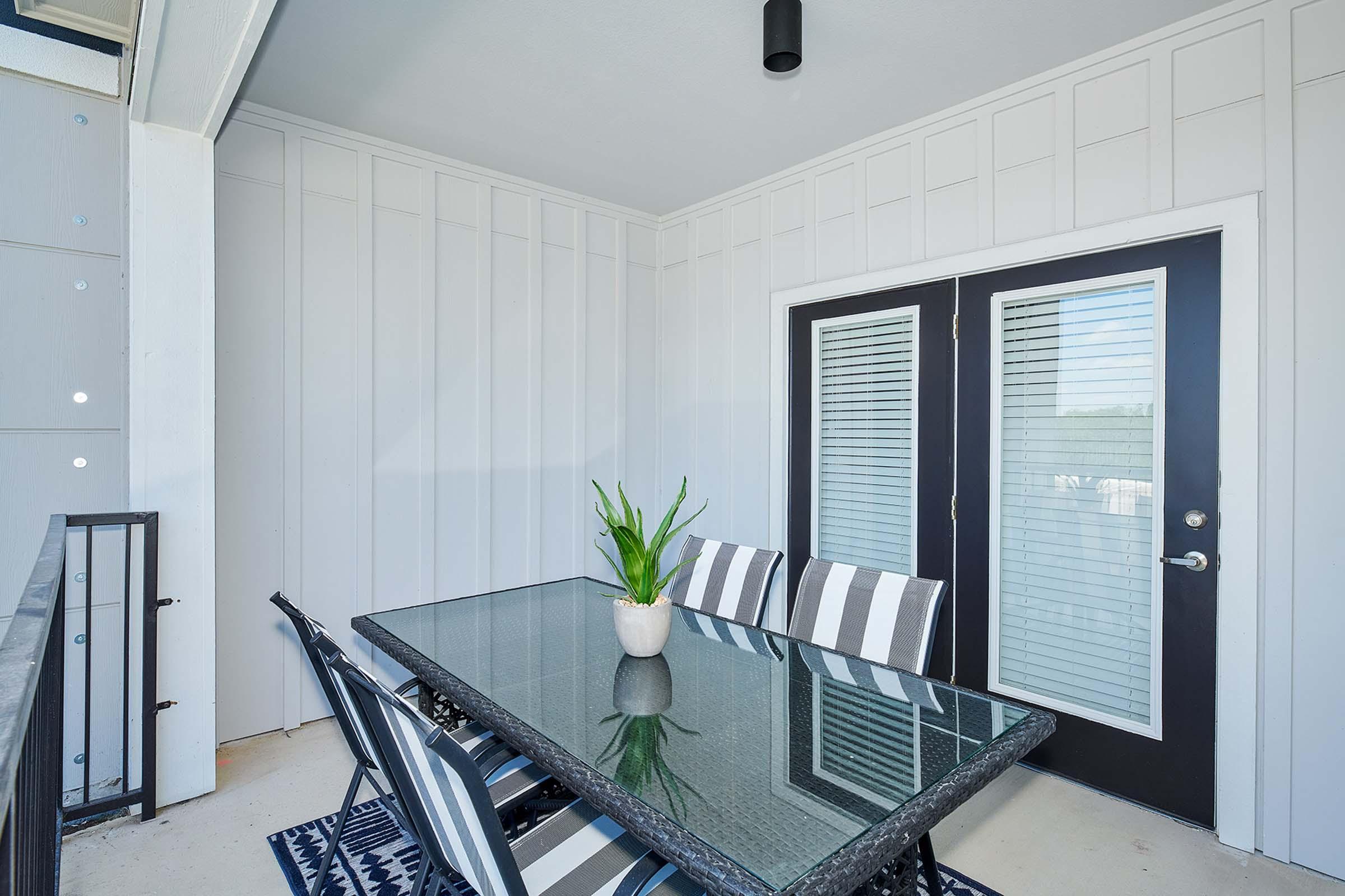 A modern balcony space featuring a glass-top dining table with eight striped chairs. The walls are painted white with a panel design, and there is a plant centerpiece on the table. Large sliding doors with blinds provide access to the outside, and the floor has a decorative blue rug.