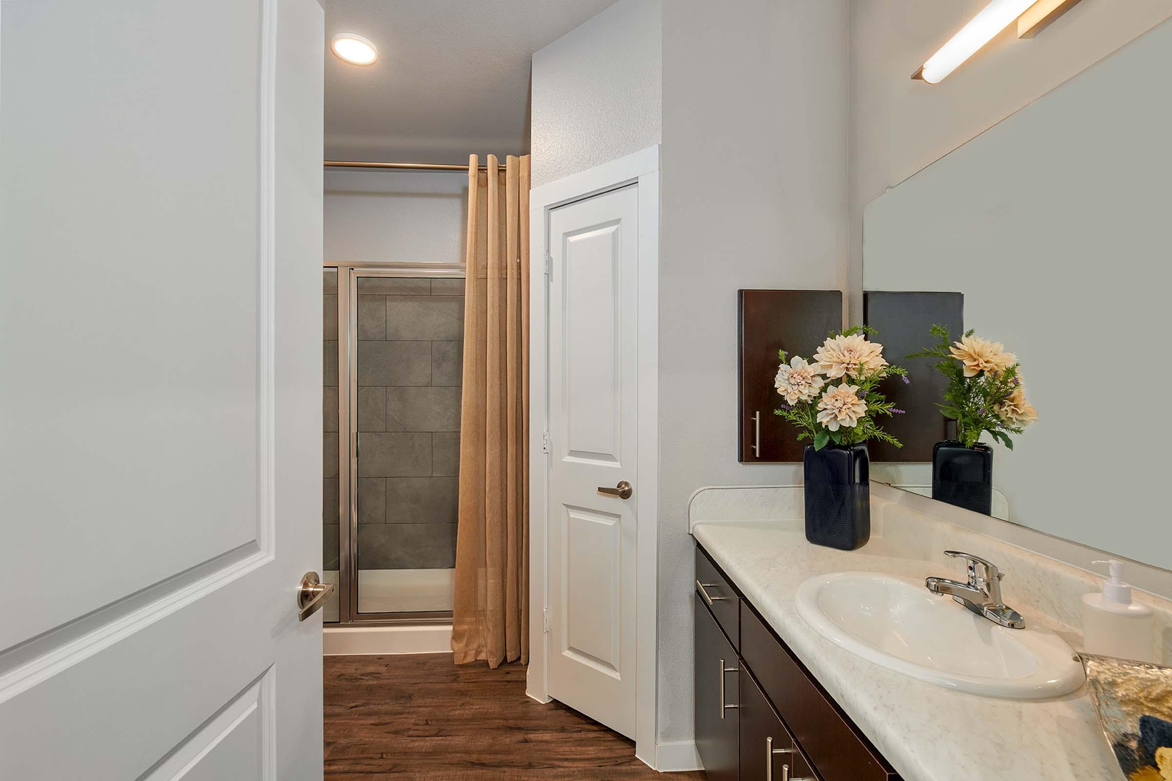 A modern bathroom featuring a white sink with dark cabinetry, a large mirror, and a glass shower enclosure with a curtain. A vase with flowers adds a decorative touch, and the floor has a wood-like finish for a warm ambiance. Natural light illuminates the space from overhead lighting.