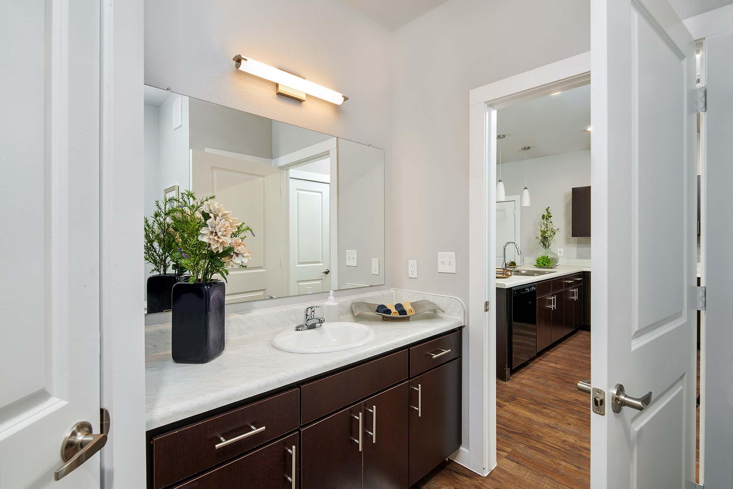 A modern bathroom featuring a sink with a dark cabinet below, a large mirror above, and a vase with flowers. Bright lighting from a wall-mounted fixture illuminates the space. The door leads to another area, showcasing a clean and stylish design with wooden flooring and minimalist decor.