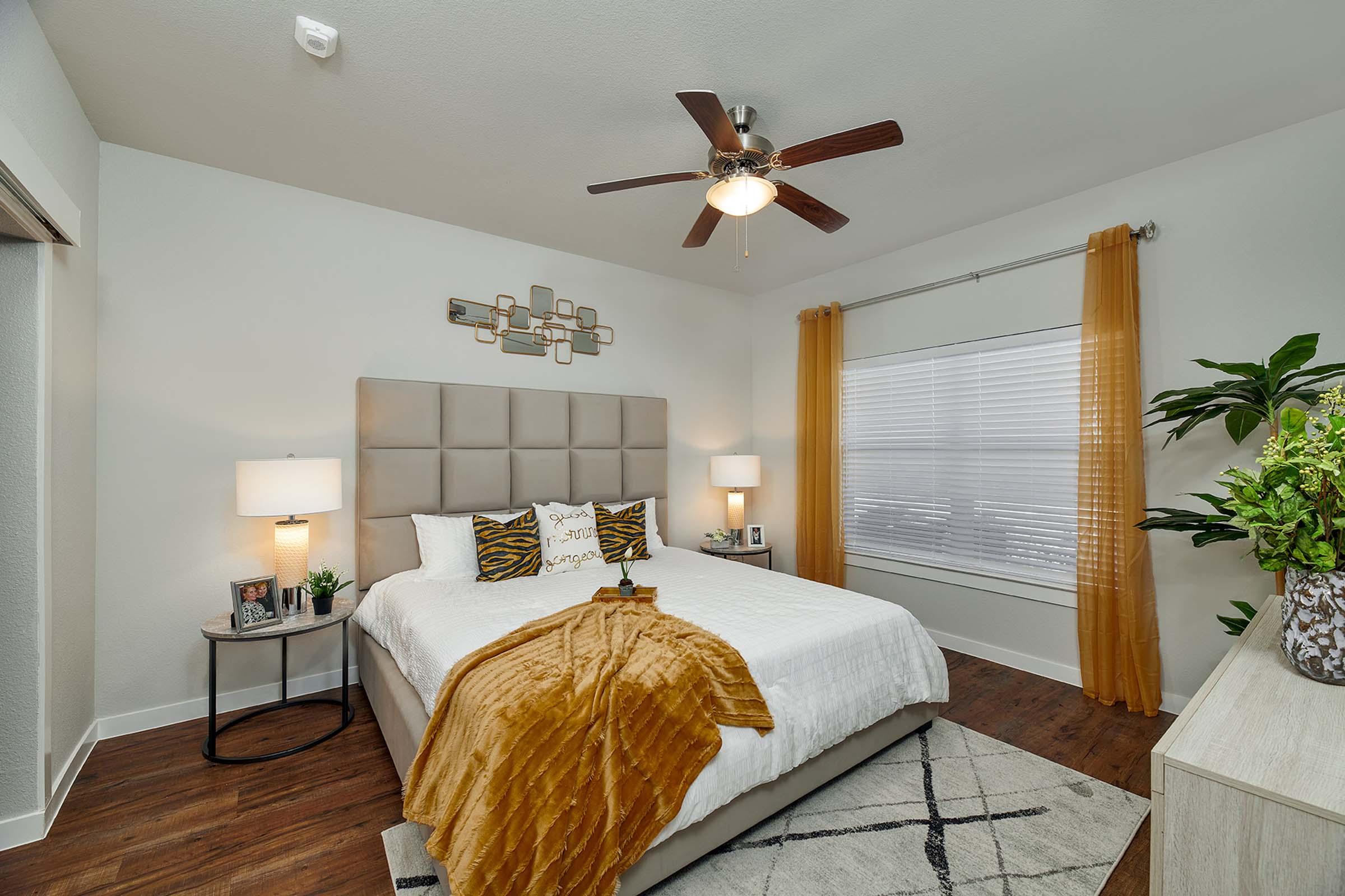 A cozy bedroom featuring a king-size bed with a light-colored comforter and decorative pillows. There are two bedside lamps on small tables, a large ceiling fan, and a stylish wall decoration. Natural light enters through a window adorned with sheer curtains, complemented by a potted plant and decorative accents.