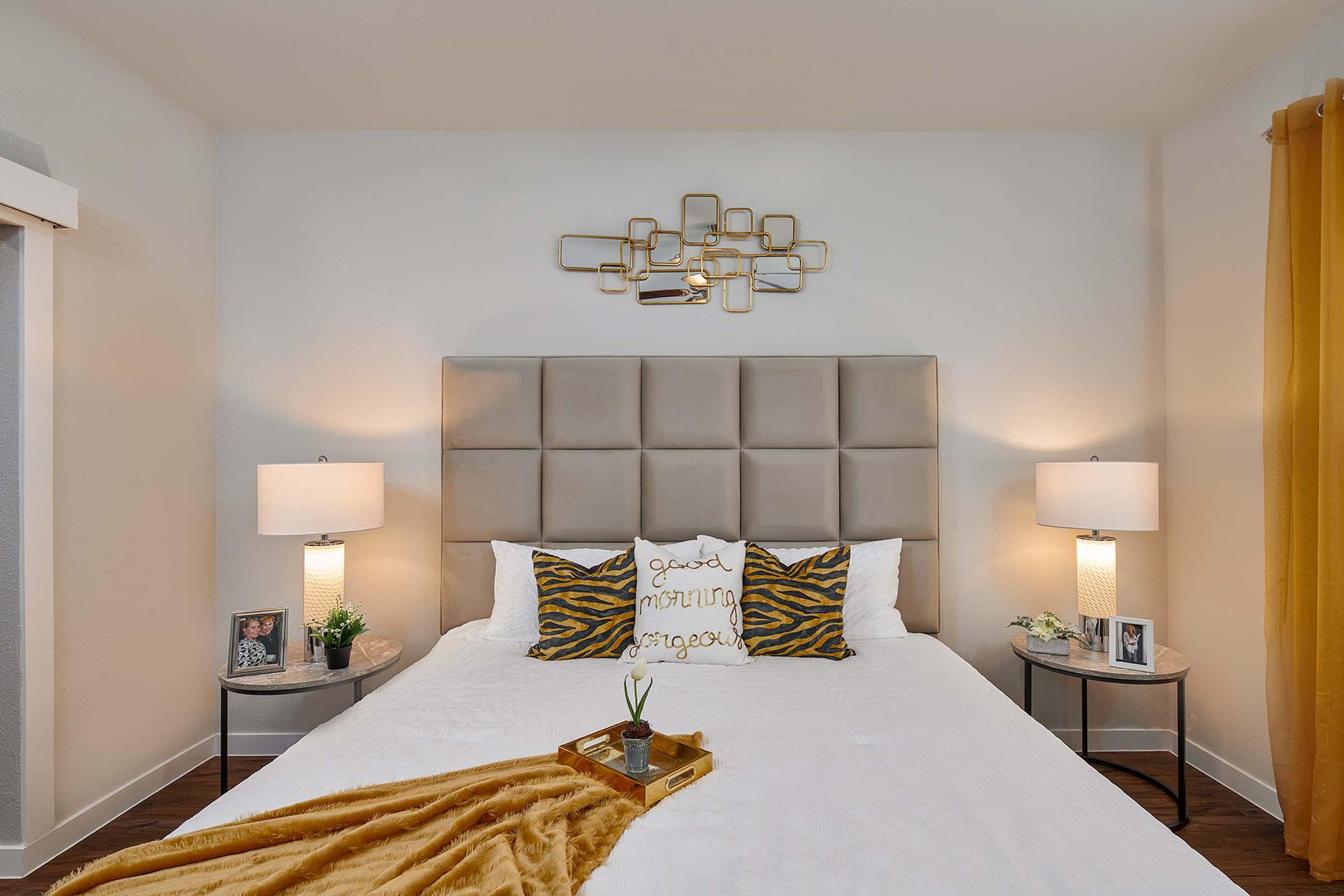 A stylish bedroom featuring a large upholstered headboard with square patterns, two bedside lamps on side tables, and decorative pillows with a zebra print. The walls are light-colored, and there is a golden wall fixture above the bed. A cozy throw blanket adds warmth to the bed, while a small green plant decorates the bedside table.