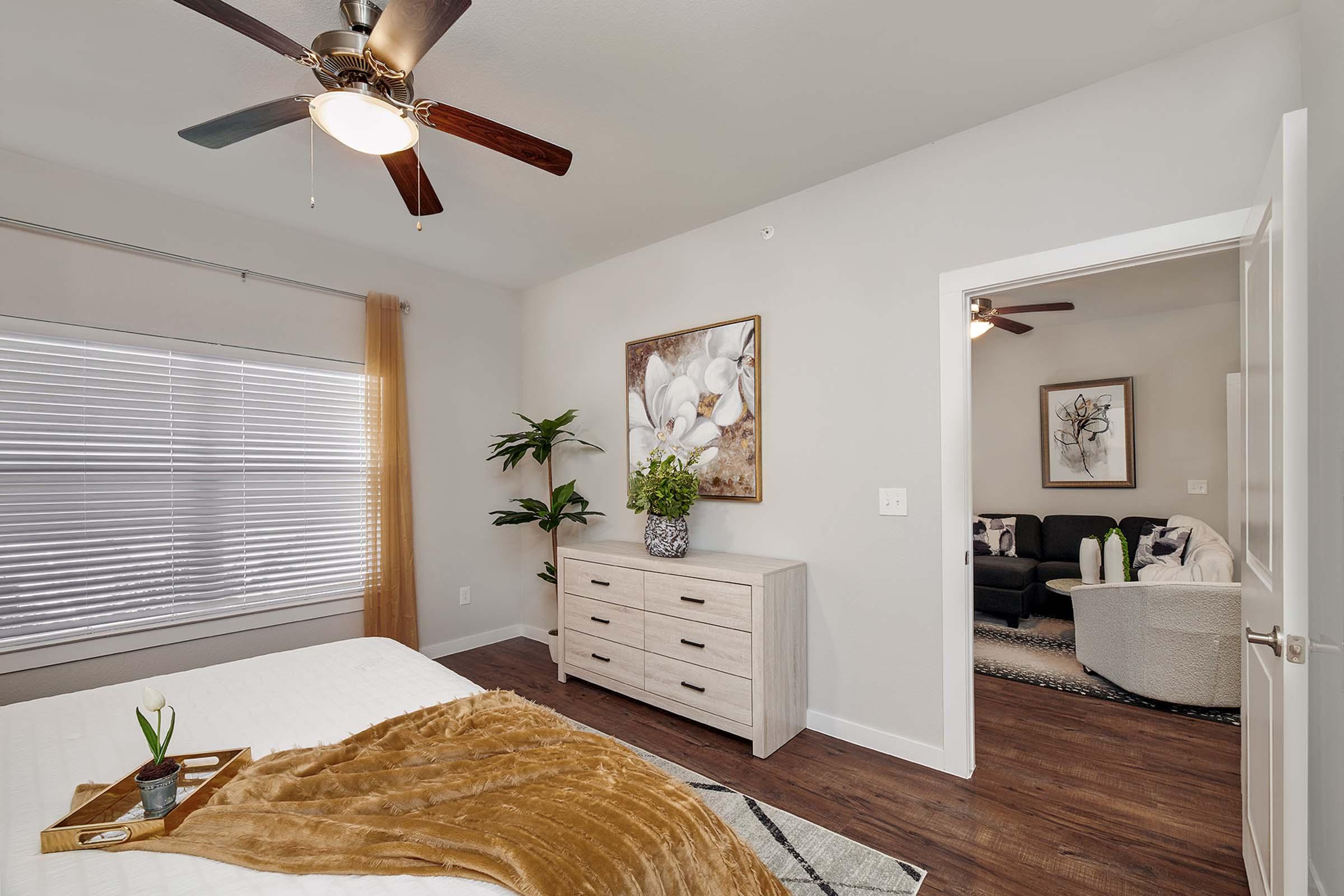 A modern bedroom featuring a bed with a soft brown blanket, a light wood dresser, and a wall art piece of flowers. A window with blinds allows natural light, and a potted plant adds greenery. The open doorway leads to a cozy living area with a black couch and additional decorative artwork on the walls.