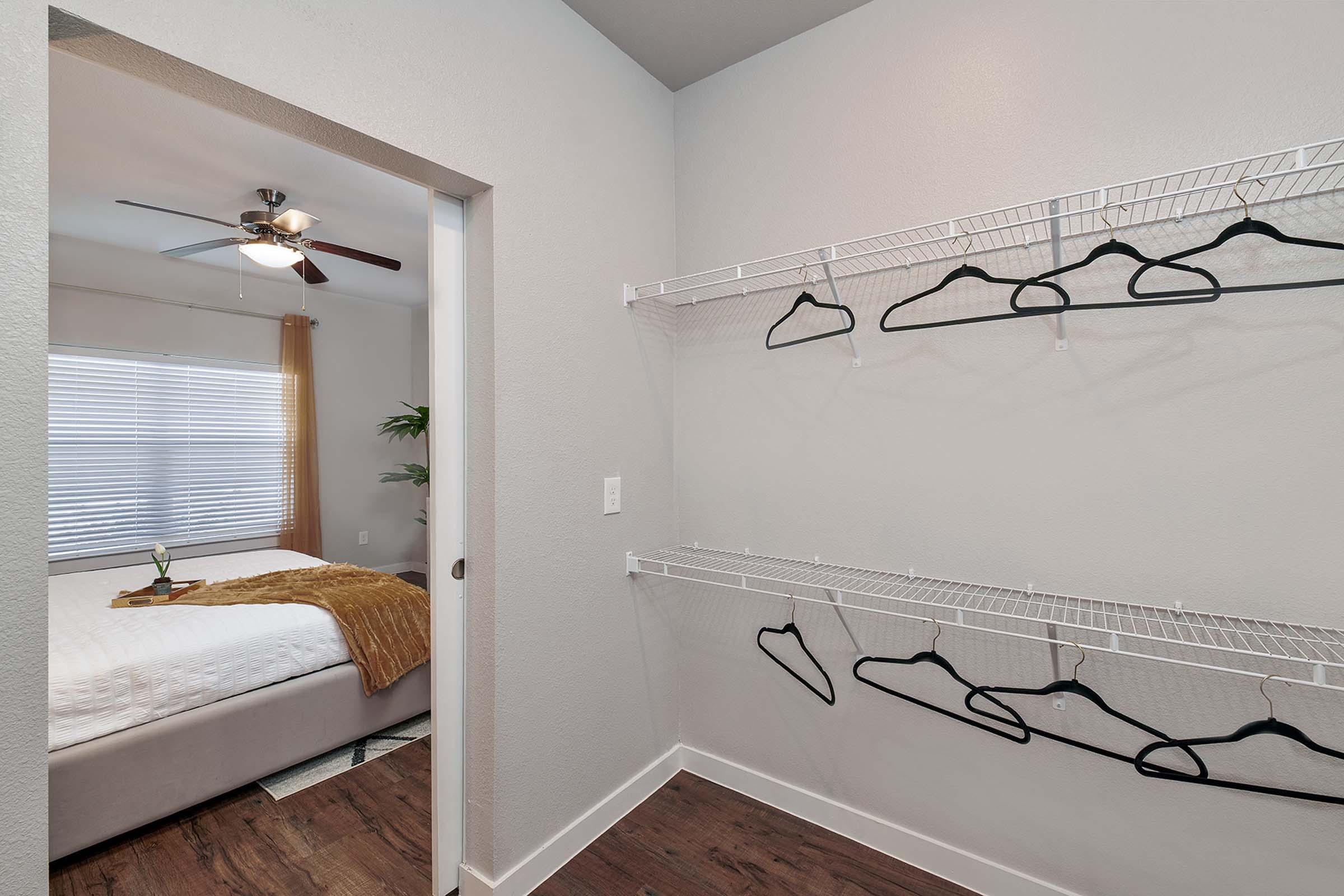 Interior view of a modern bedroom from a hallway. The room features a bed with a cozy blanket, a ceiling fan, and bright natural light coming through a window with simple curtains. To the side, there are wire shelves with black hangers, ideal for storage or organization.