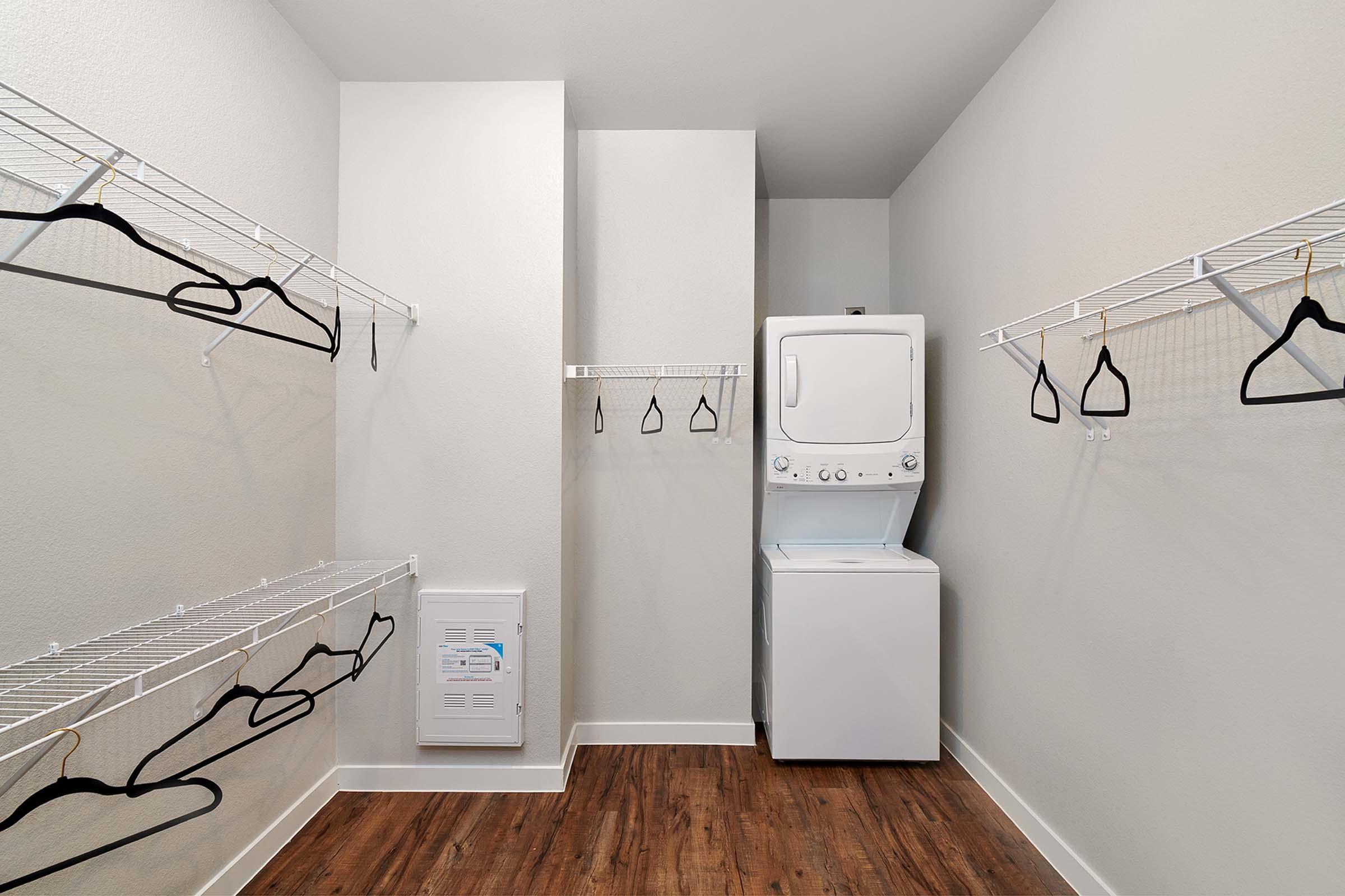 A clean, organized laundry room featuring a stacked washer and dryer unit. Two wire shelving units with several empty black hangers are mounted on the walls, providing storage space. The room has light-colored walls and a wood laminate floor, creating a bright and inviting atmosphere.