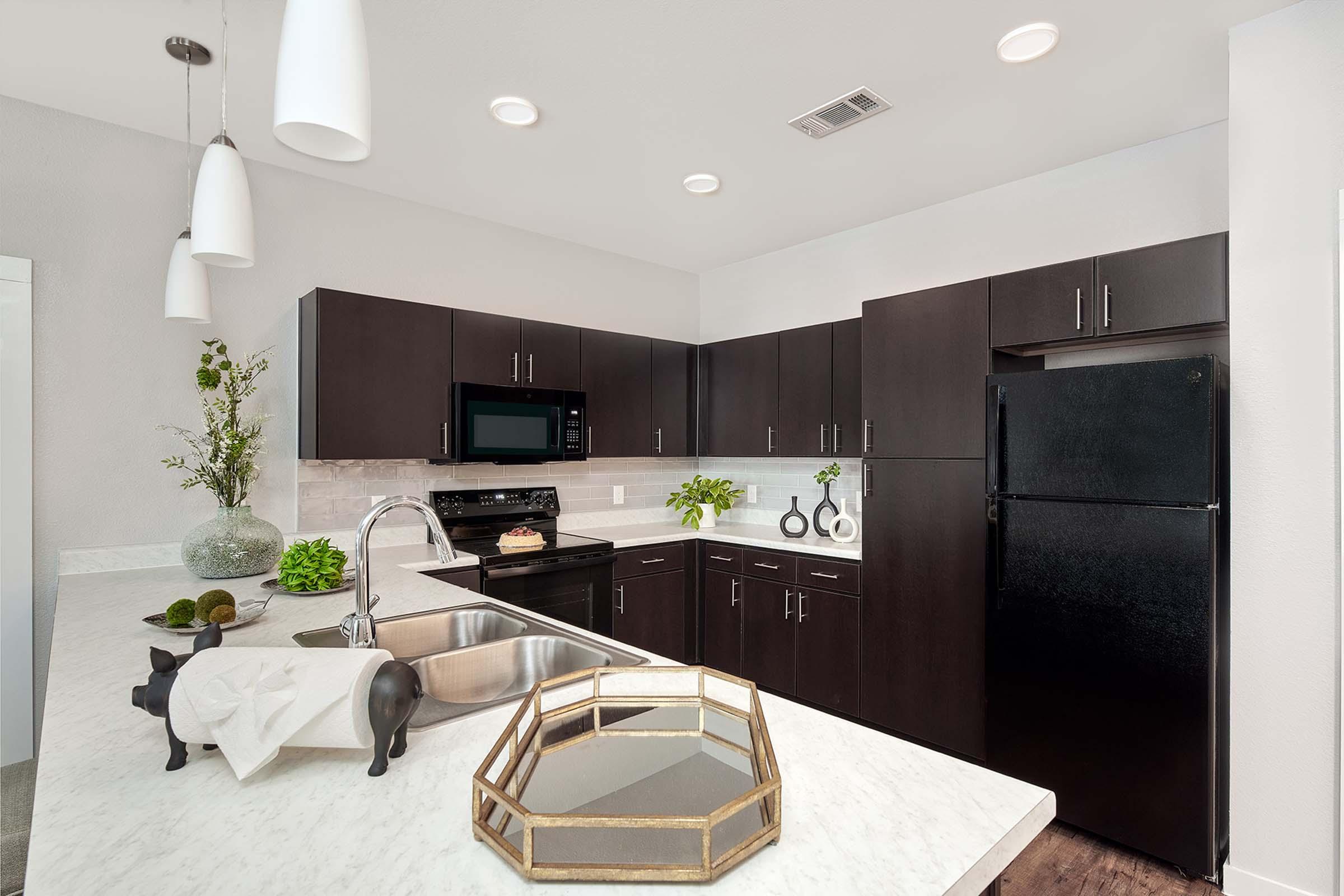 A modern kitchen with dark cabinets and a light marble countertop. There is a black refrigerator, a microwave, and an elegant centerpiece on the counter. The space is well-lit with recessed lighting and features decorative plants for a fresh touch.