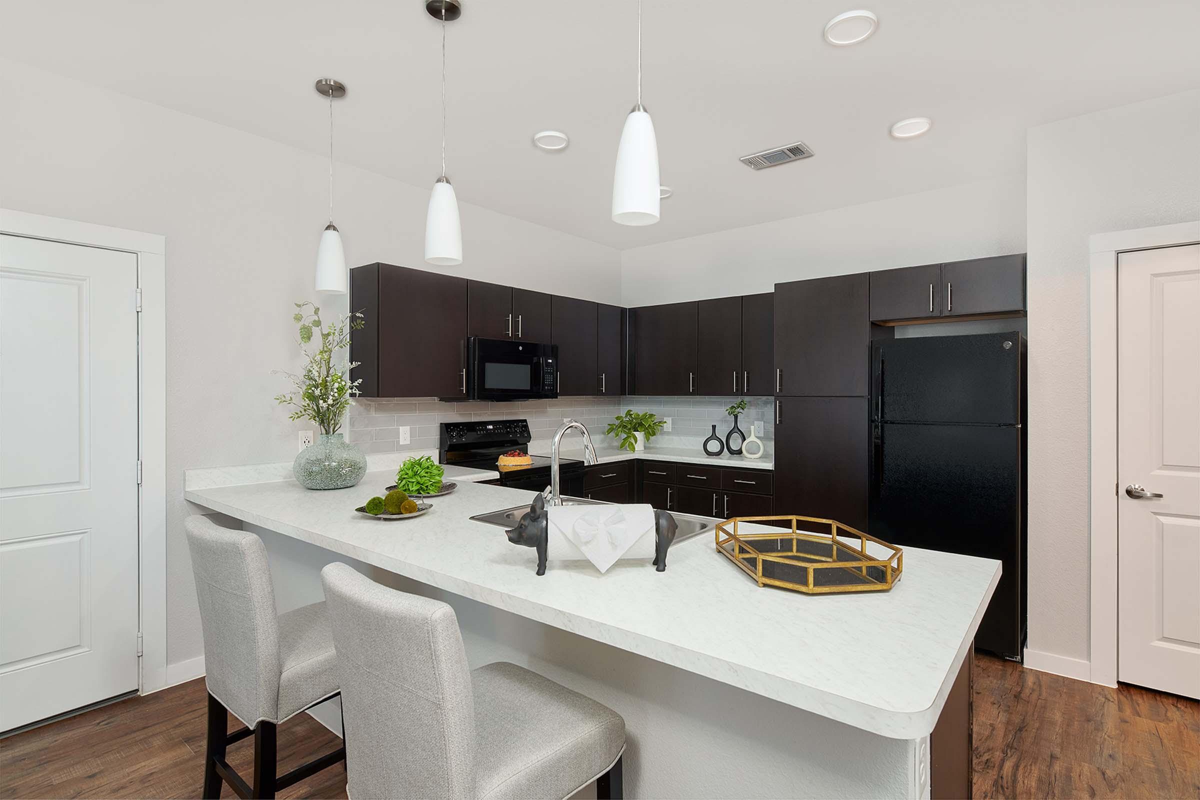 Modern kitchen featuring dark wood cabinets, stainless steel appliances, and a white countertop. A large island with bar stools is positioned in the foreground, accompanied by decorative items like plants and a serving tray. The space is well-lit with pendant lights and has a minimalist design.