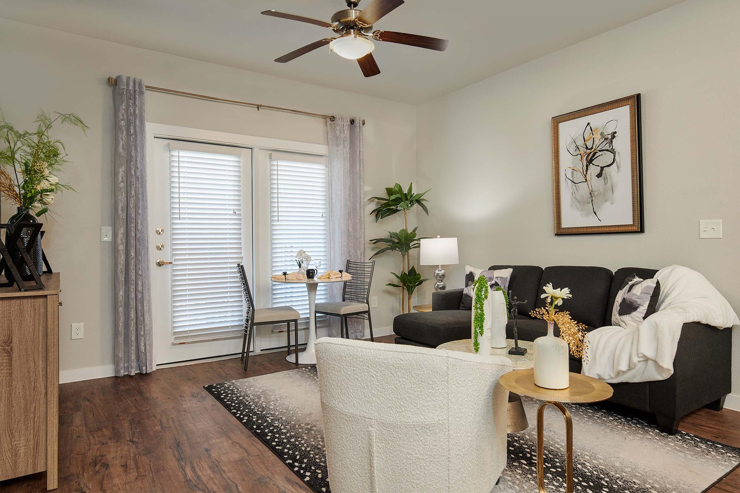 A cozy living room featuring a dark gray sofa, white armchair, and wooden coffee table. A small dining area with two chairs and a table is positioned near glass doors. Decorative plants, a lamp, and artwork add elegance to the space, which has light-colored walls and dark wood flooring.