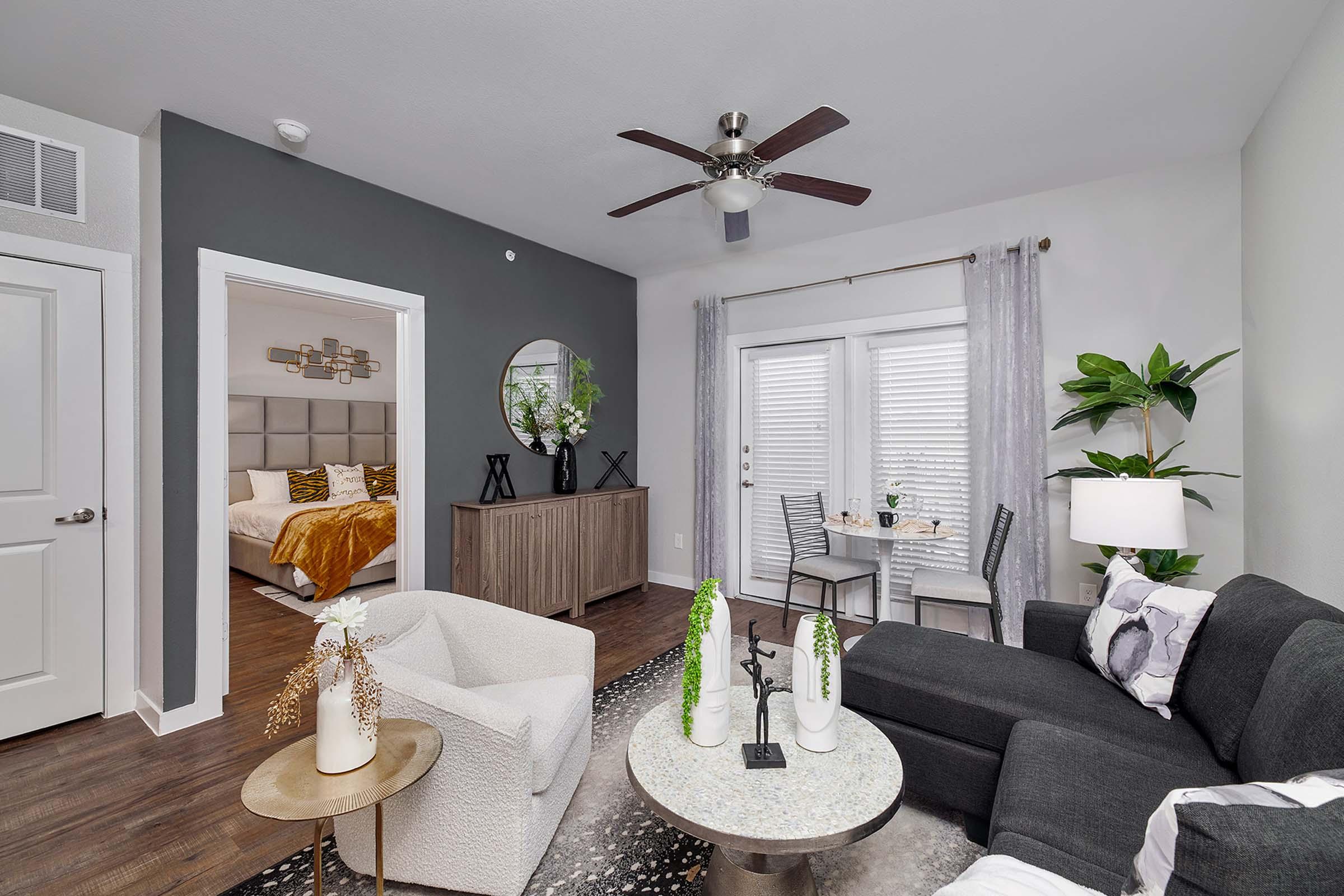 A modern living room featuring a dark green accent wall, a black sectional sofa, and a round coffee table with decorative plants. In the background, there's a dining area with a small table and two chairs. A bedroom is visible through an open doorway, showcasing a bed with decorative pillows.