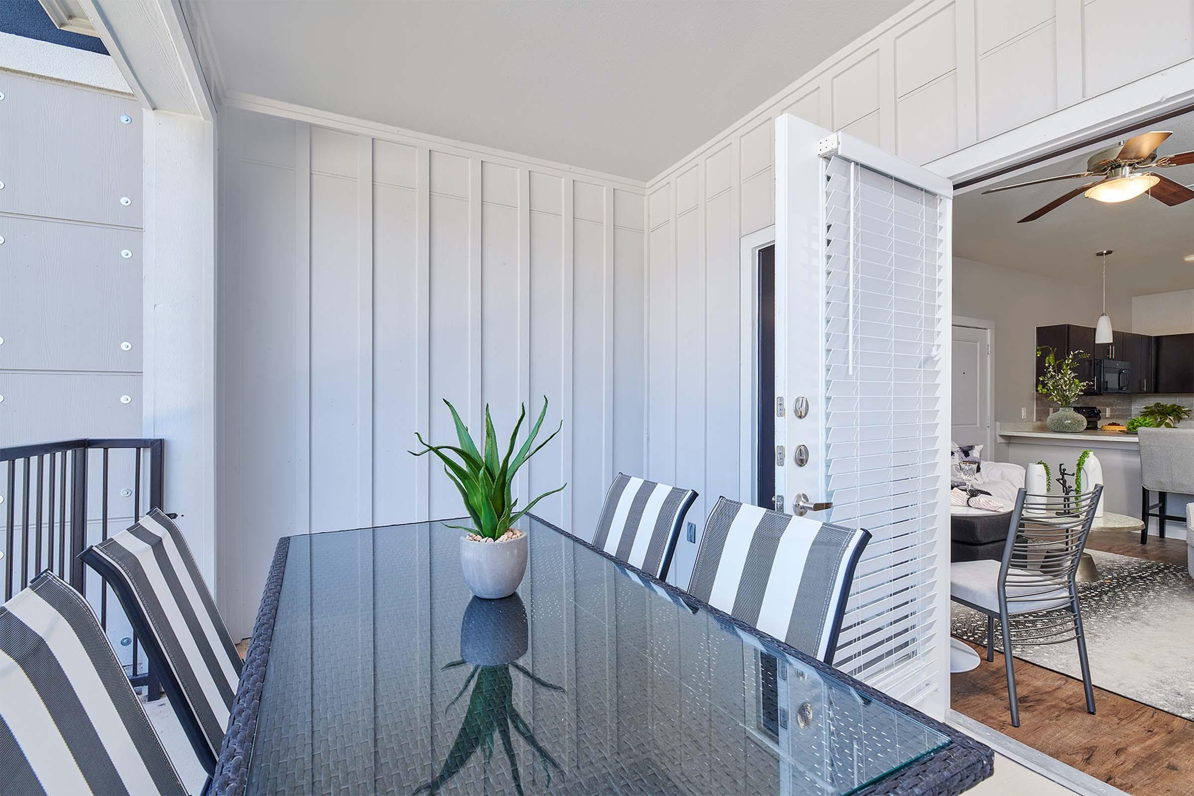 A modern dining area featuring a glass-top table surrounded by striped chairs. There is a potted plant in the center of the table. In the background, a set of open doors leads to a well-lit kitchen area, with a ceiling fan visible above and a rug on the floor. Bright and inviting decor.