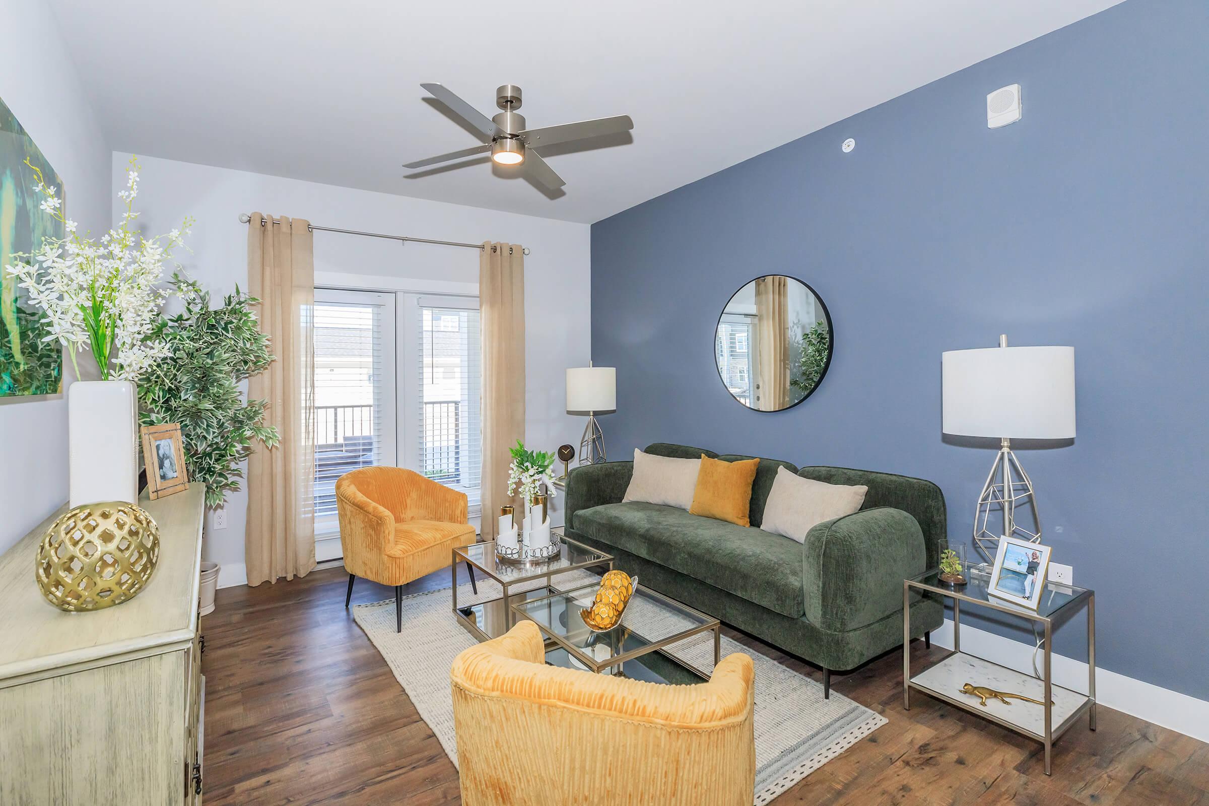 A modern living room featuring a green sofa, two orange armchairs, a glass coffee table, and decorative plants. The walls are a calming blue, and a large round mirror reflects light. Natural light streams in through sliding glass doors, complemented by elegant curtains and stylish lighting fixtures.