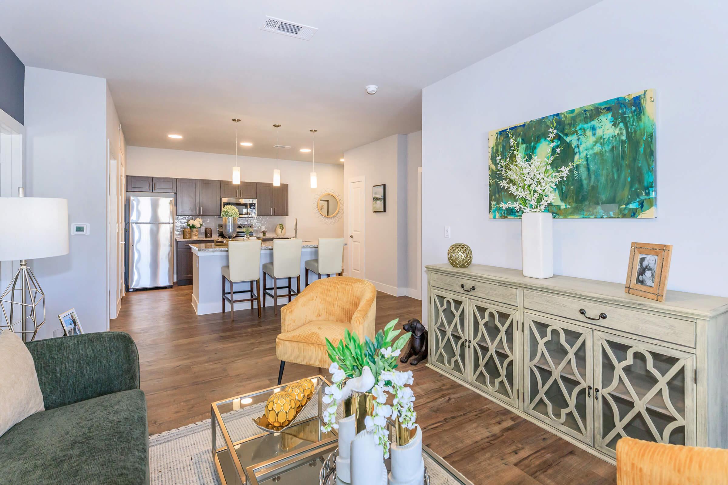 A modern living room and kitchen area featuring a green sofa, decorative pillows, and a light-colored sideboard. The kitchen is visible in the background with bar stools, stainless steel appliances, and stylish decor. A painting and a vase with flowers add a touch of elegance to the space.