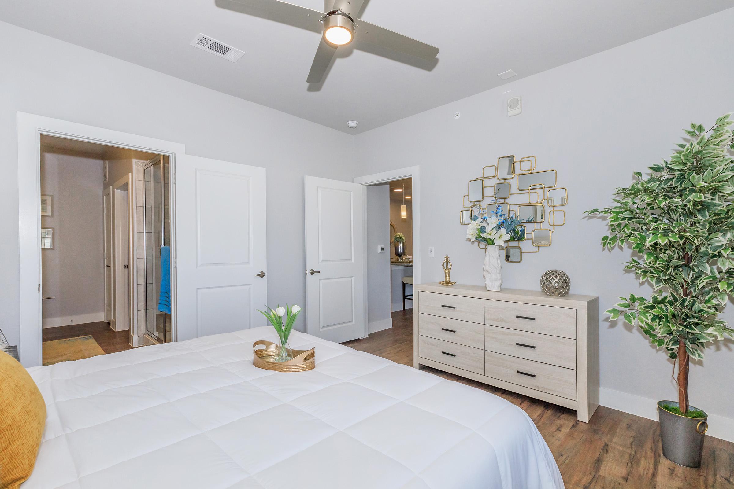 A well-decorated bedroom featuring a bed with white linens and a yellow accent pillow. A wooden dresser with decorative items and a potted plant is visible. The room has light gray walls, a ceiling fan, and a doorway leading to a bathroom, creating a bright and inviting atmosphere.