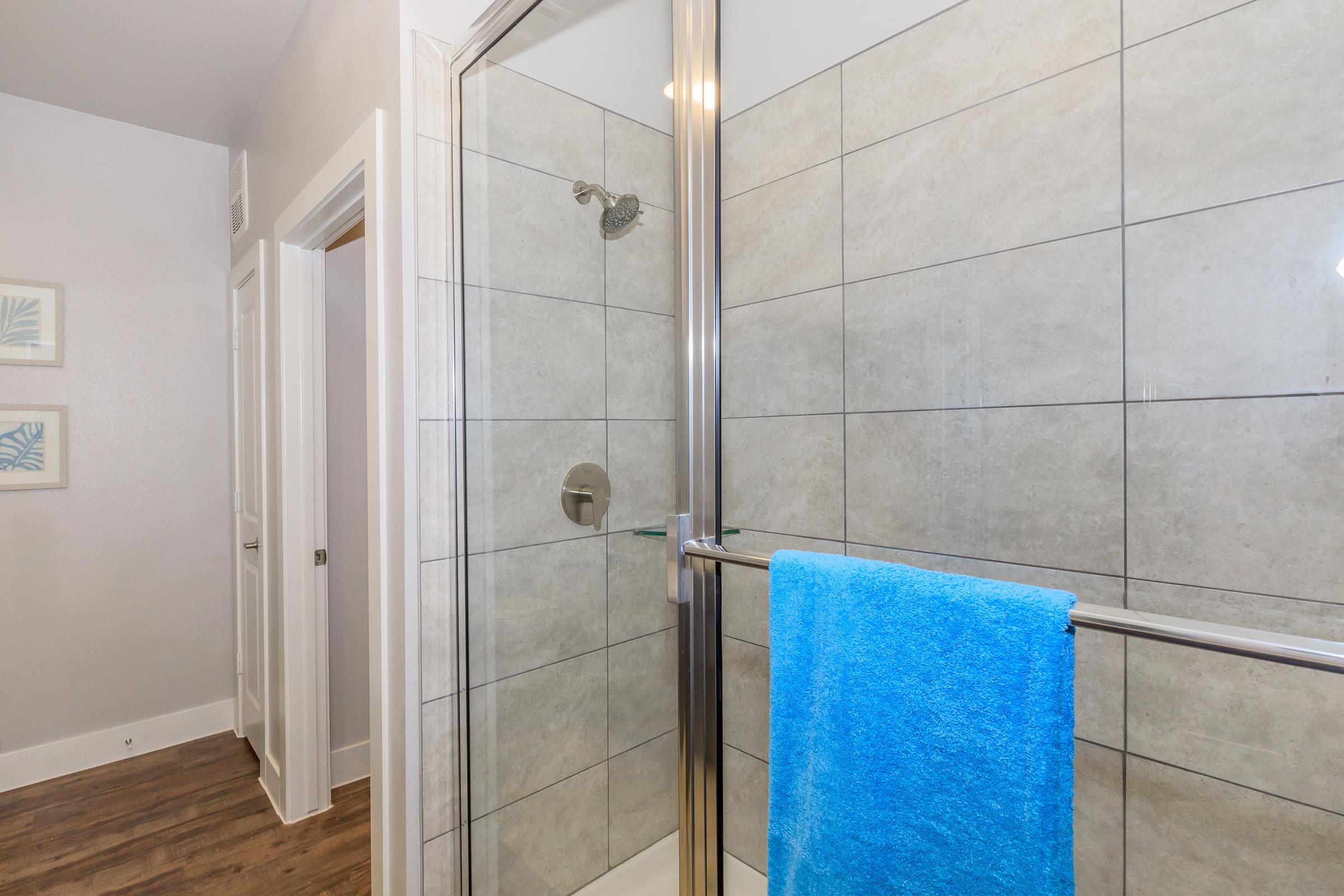 A modern bathroom shower with a glass door, featuring tiled walls in neutral colors. A chrome towel rail holds a bright blue towel. The floor is wooden, and a white door can be seen in the background. The overall aesthetic is clean and contemporary.