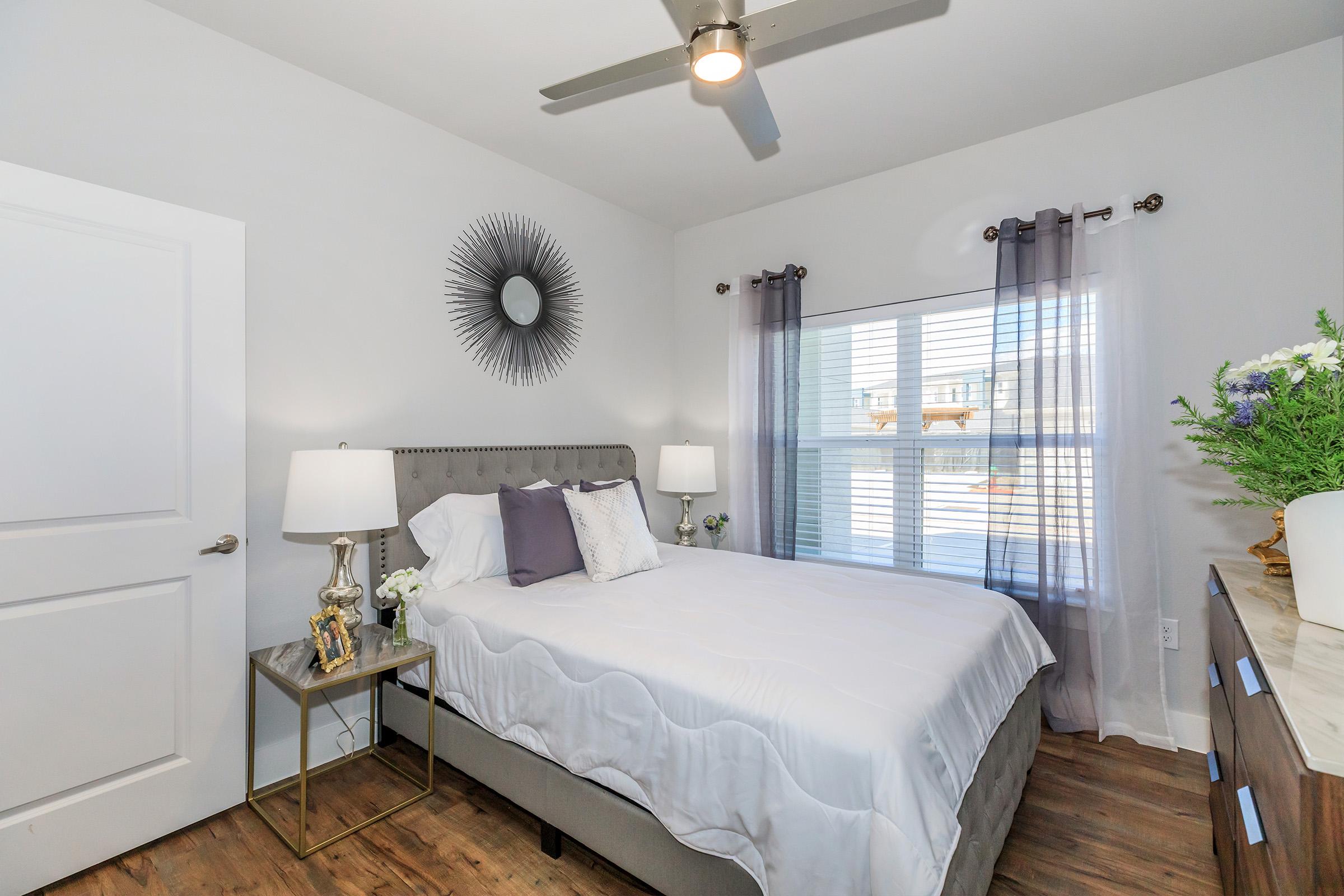 Bright and airy bedroom featuring a queen-sized bed with white linens, a decorative headboard, and two bedside lamps. The room has large windows with sheer curtains, a stylish wall mirror, and a minimalist dresser with decorative flowers, creating a cozy and inviting atmosphere.