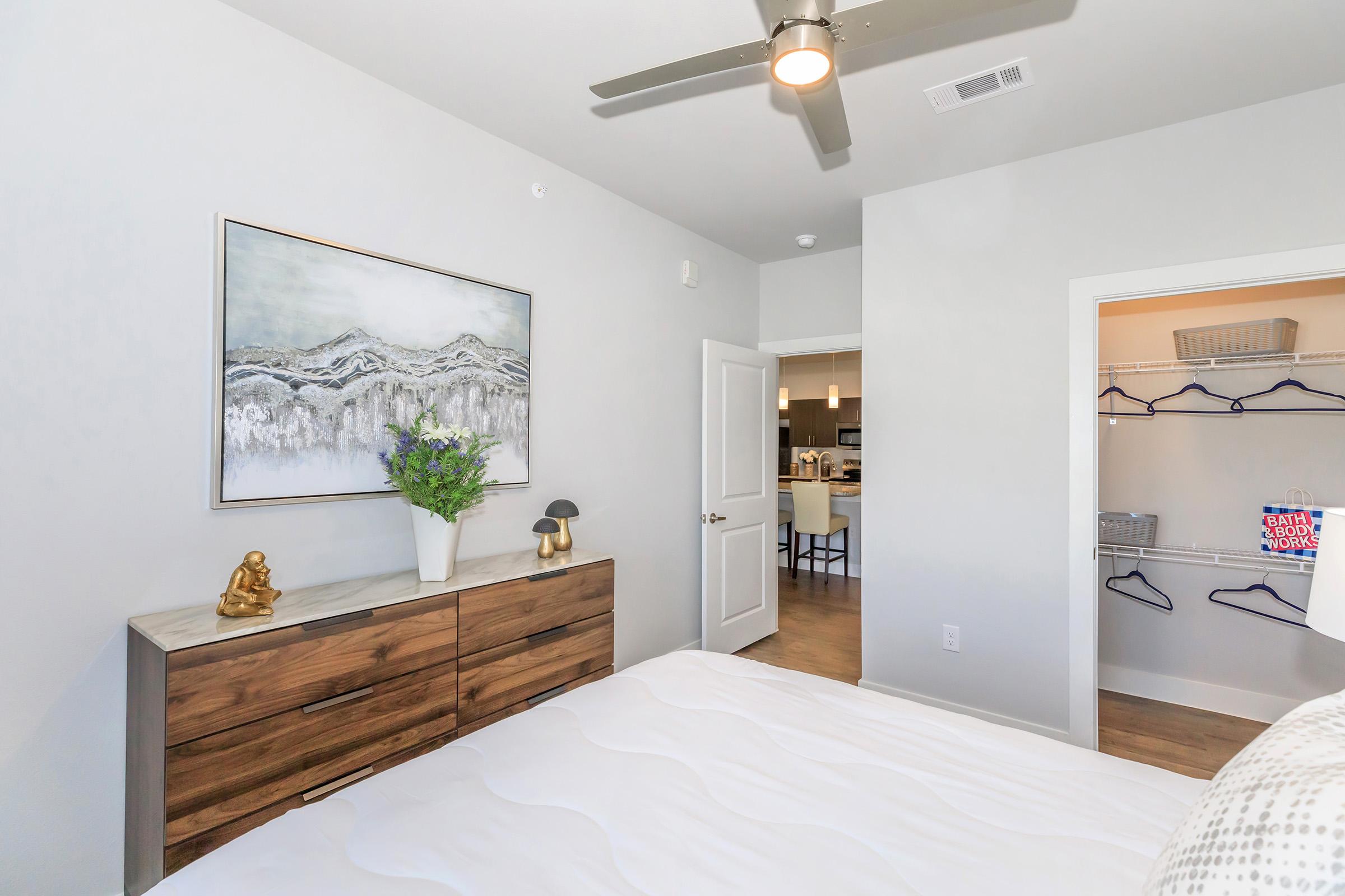 A modern bedroom featuring a neutral color palette, a wooden dresser with decorative items, a large abstract painting on the wall, a ceiling fan, and an open closet space. Through the door, a glimpse of a kitchen area can be seen.