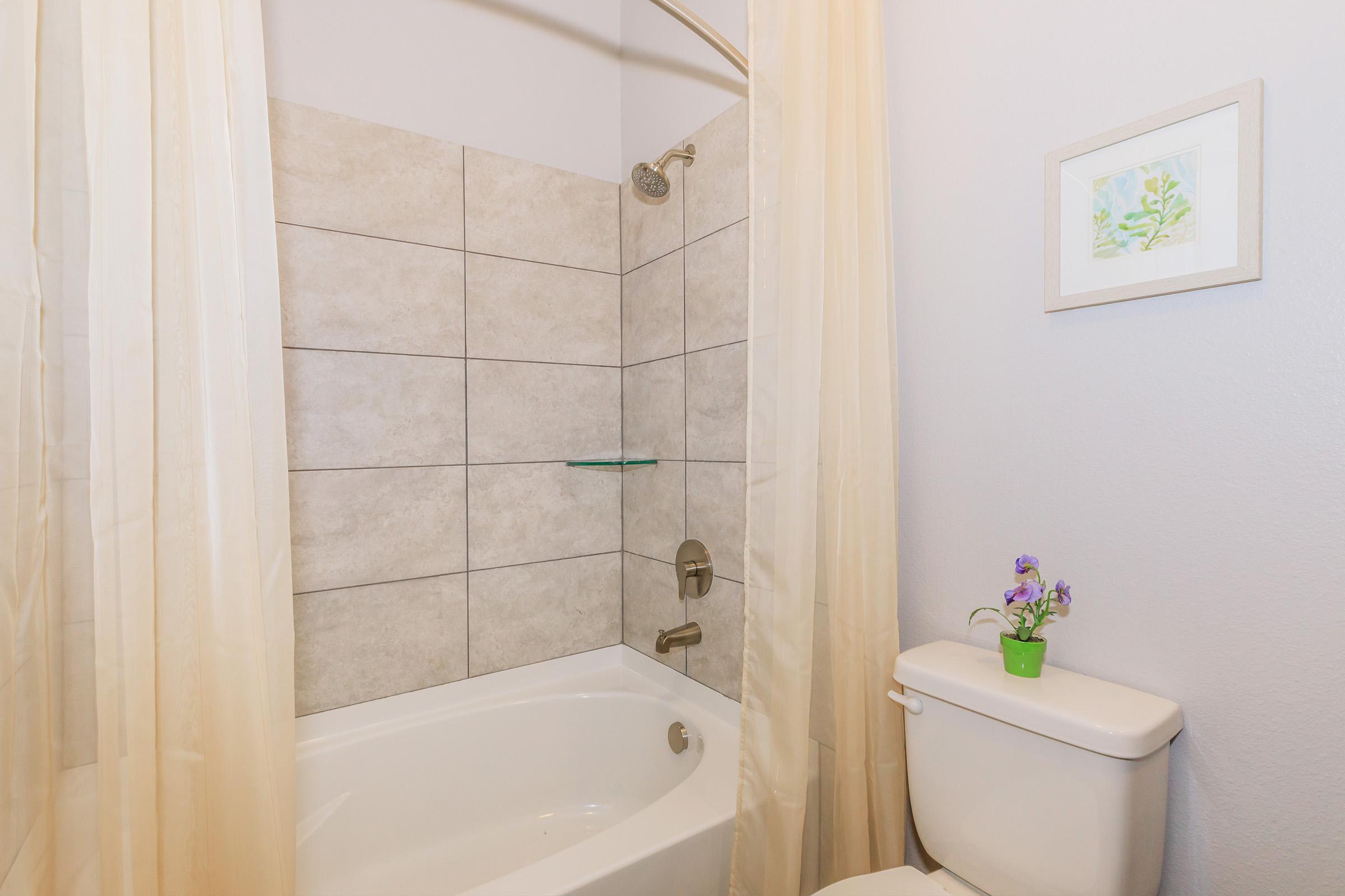 A bathroom featuring a bathtub with a shower curtain, light gray tile walls, and a glass shelf. There is a modern, white toilet and a small potted plant with purple flowers on top. A framed piece of art is hung on the wall, adding a decorative touch to the space.