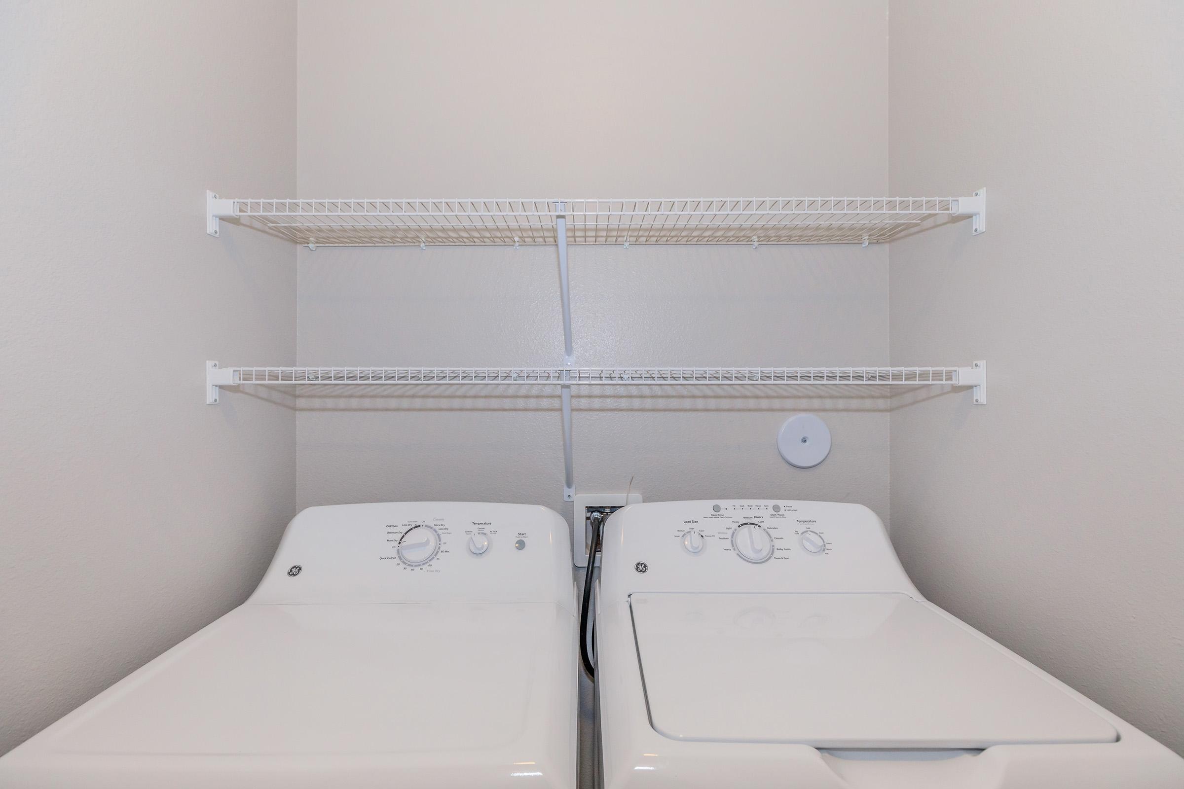 A clean laundry room featuring a stacked washer and dryer side by side, with a set of white wire shelves mounted above them. The walls are a light color, creating a bright and organized space suitable for laundry tasks.